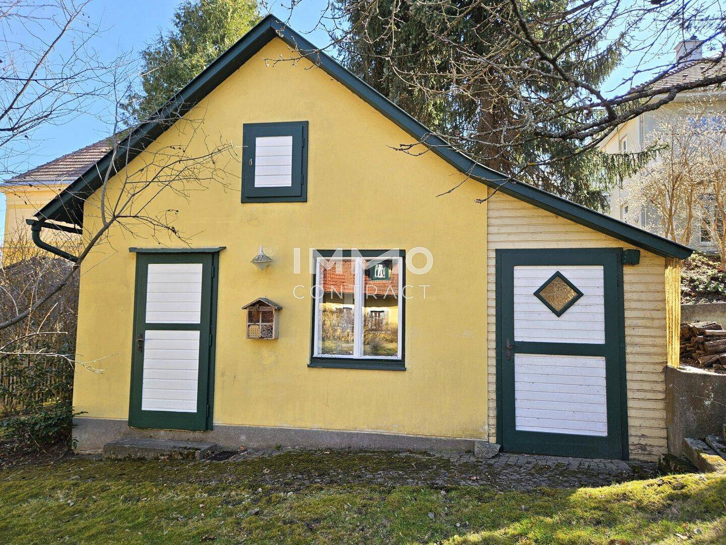Kleines Nebengebäude mit gelber Fassade und grünen Türen im Gartenbereich.