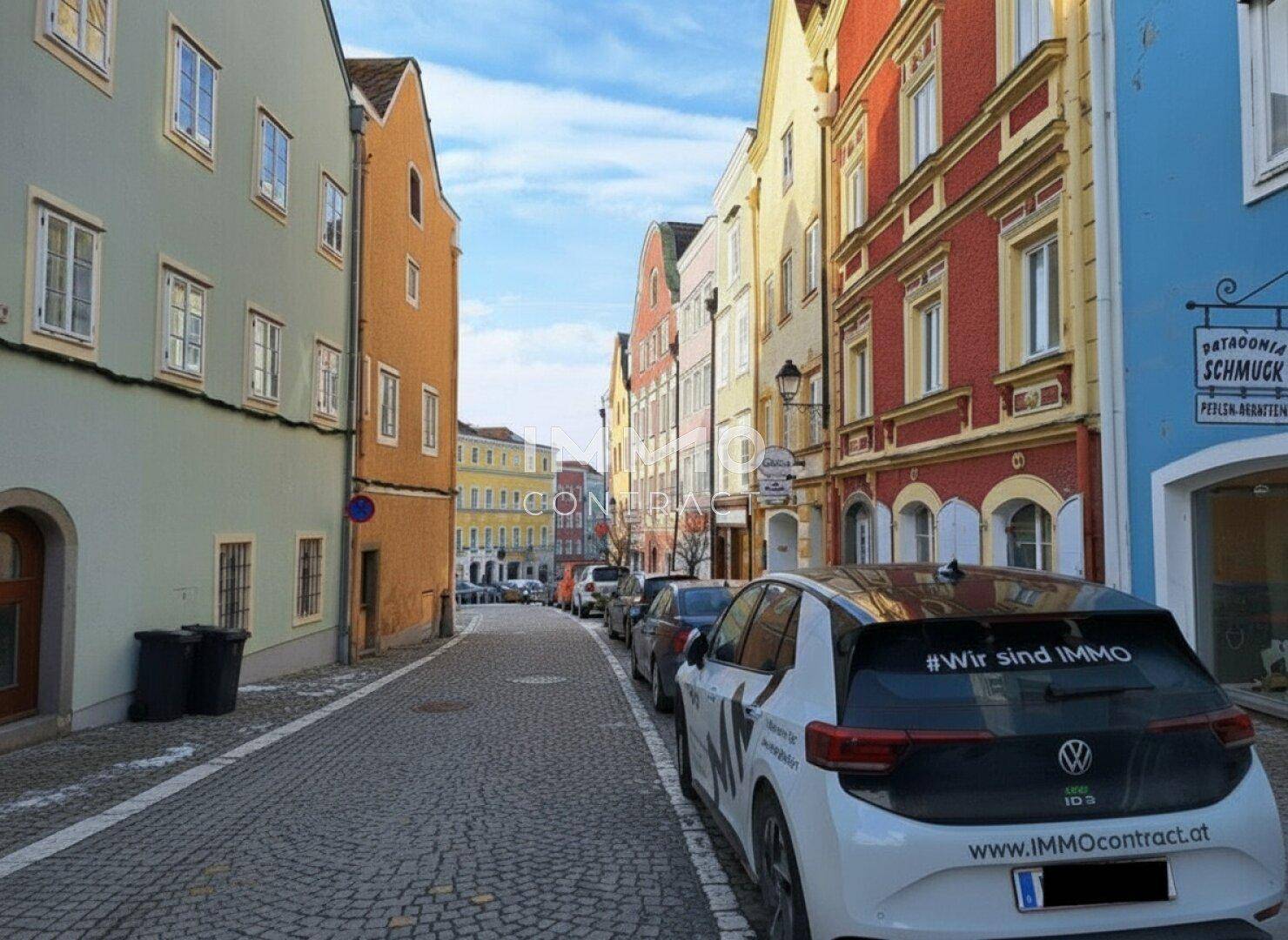 Kopfsteingepflasterte Gasse mit bunten historischen Gebäuden und geparkten Autos.