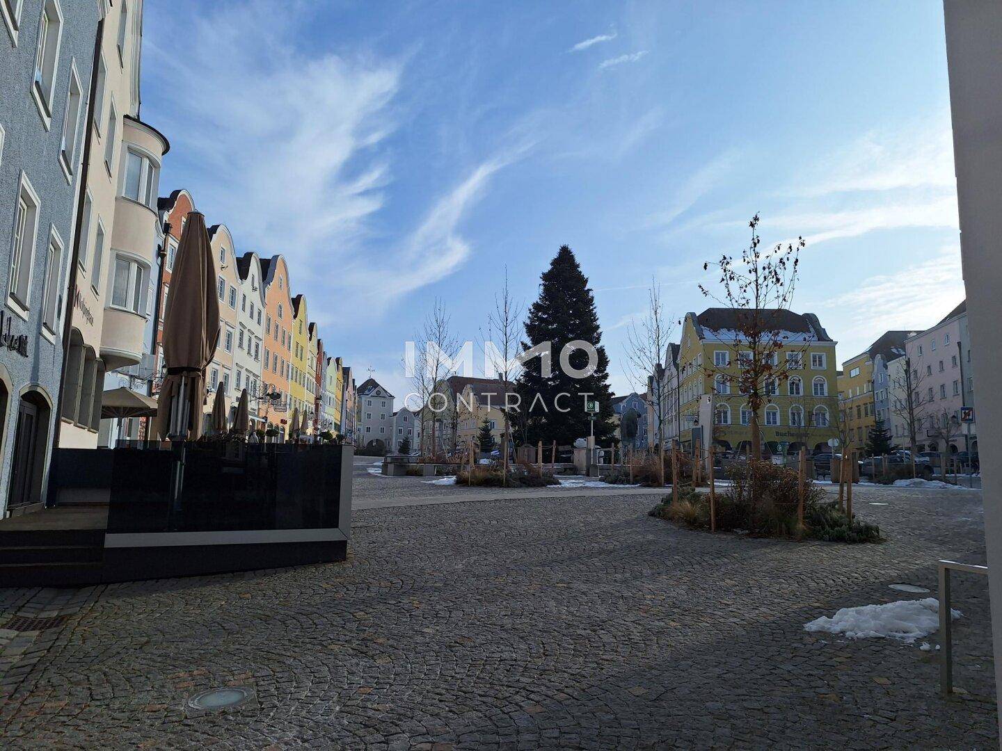 Öffentlicher Platz mit Kopfsteinpflaster, Bäumen und historischen Gebäuden unter blauem Himmel.
