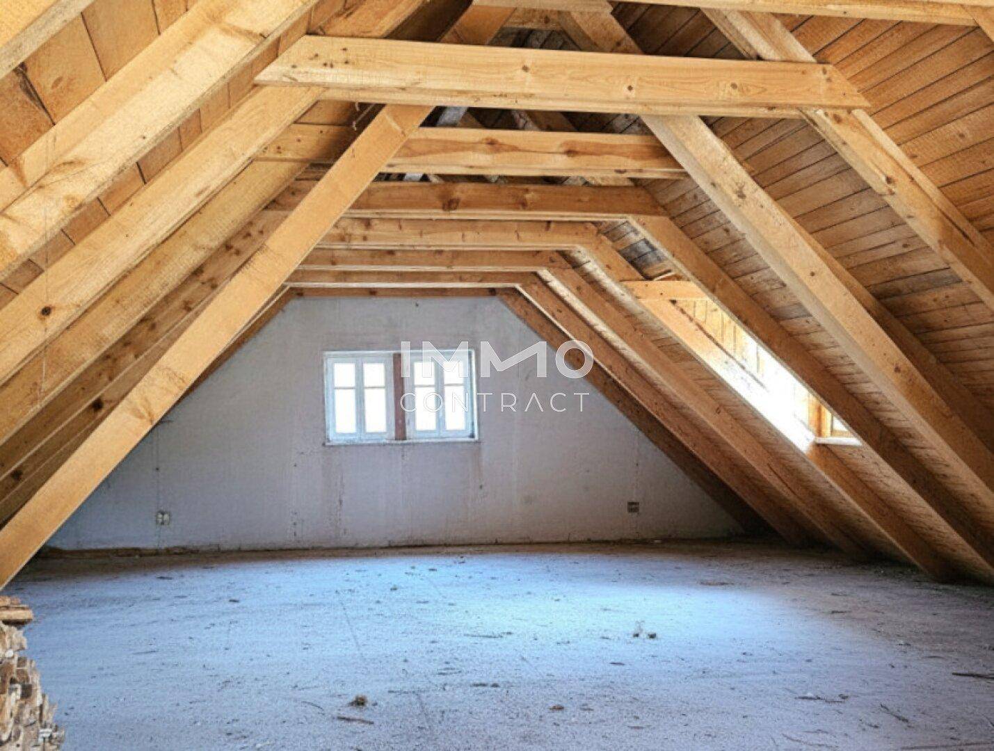 Unfertiger Dachboden mit sichtbaren Holzbalken, schrägen Wänden und Fenstern für zukünftigen Ausbau.