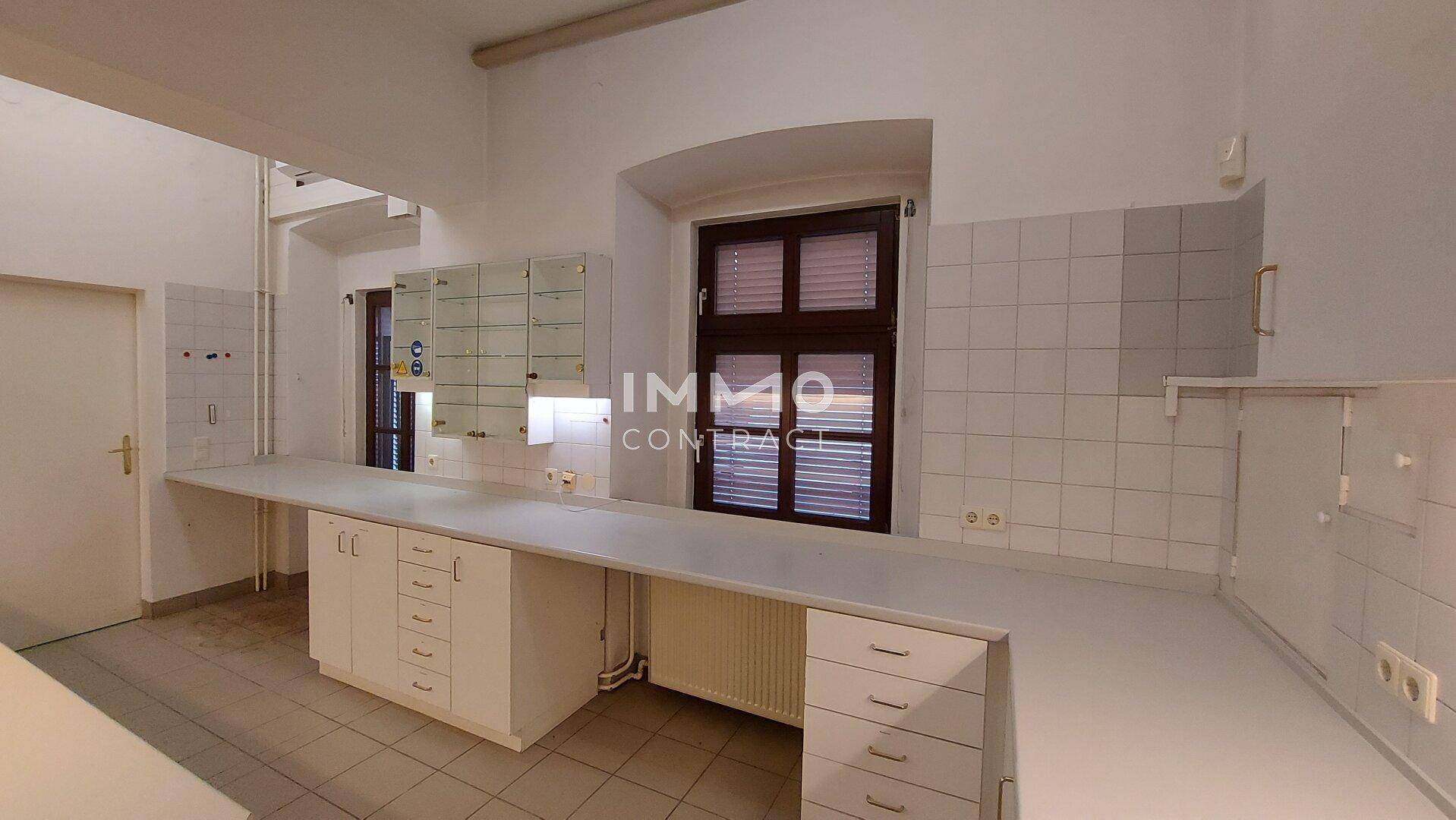 Helle Küche mit weißen Schränken, Fliesenboden und einem Fenster mit Heizkörper.