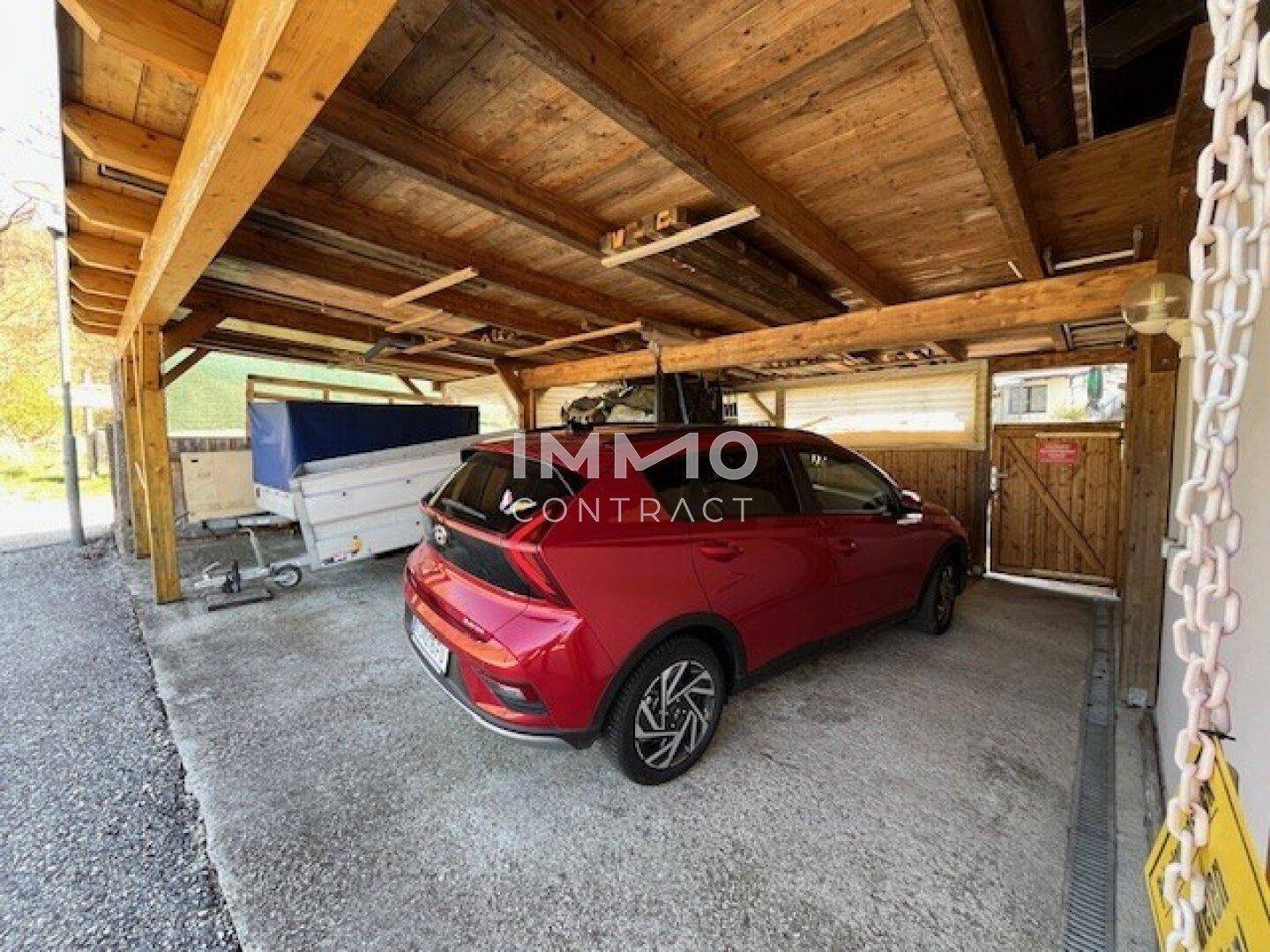 Überdachter Carport aus Holz mit Kiesboden, bietet Schutz für ein Fahrzeug und einen Anhänger.