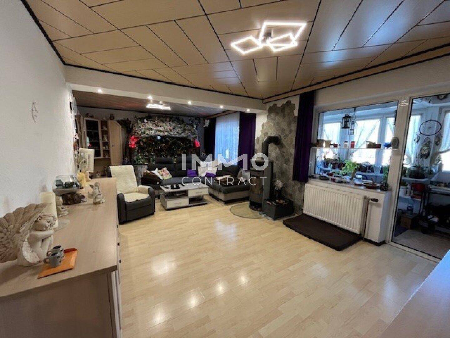 Geräumiges Wohnzimmer mit Holzboden, Kaminofen und großen Fenstern, die viel Licht hereinlassen.