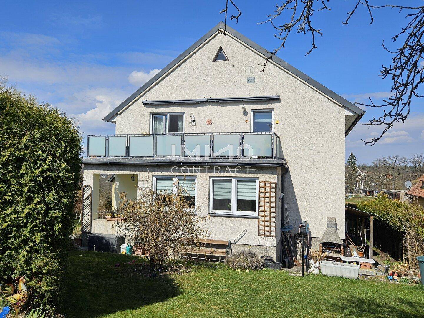 Rückansicht des Hauses mit Garten, Balkon und Außenbereich mit Grillstelle unter blauem Himmel.