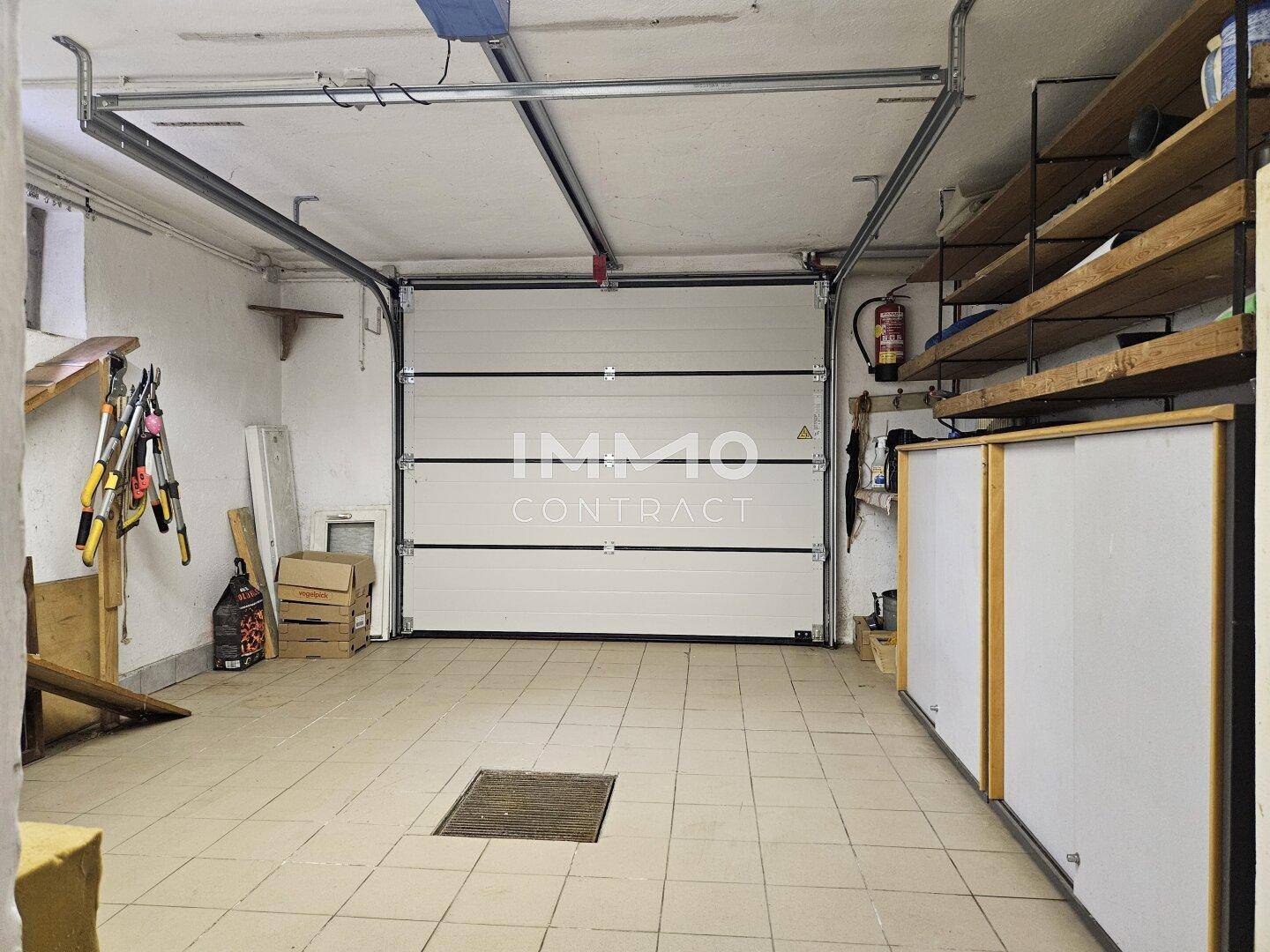 Geräumige Garage mit Fliesenboden, Sektionaltor und praktischen Regalen für Stauraum.