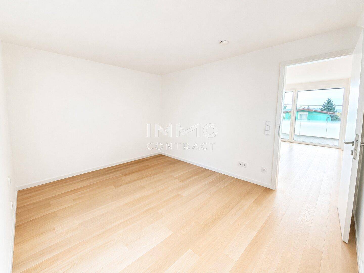 Leeres Zimmer mit hellem Holzboden und Zugang zu einem weiteren Raum mit Terrasse.