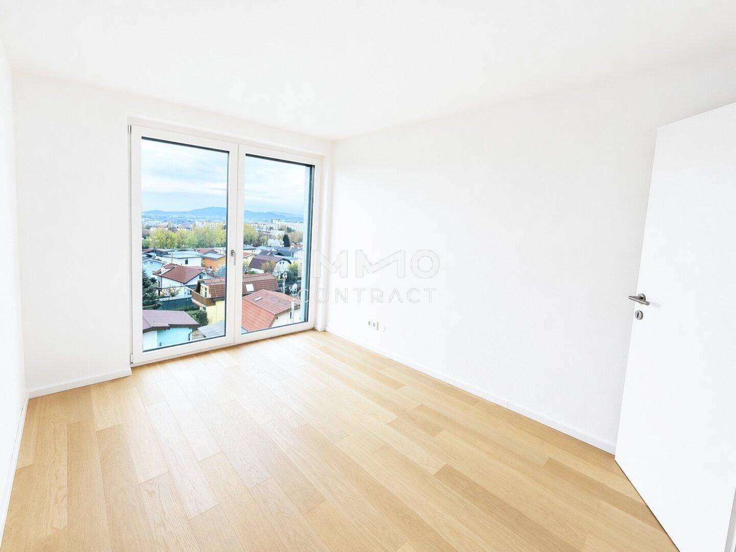 Helles Schlafzimmer mit Parkettboden und großem Fenster, das einen Blick ins Grüne bietet.
