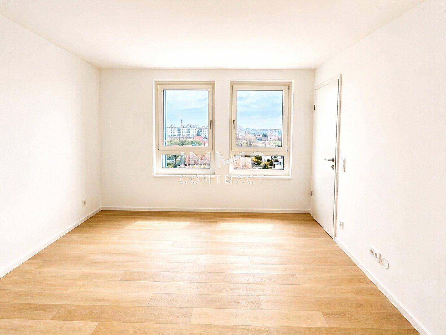 Helles, leeres Zimmer mit zwei Fenstern, Holzboden und Blick auf die Umgebung.