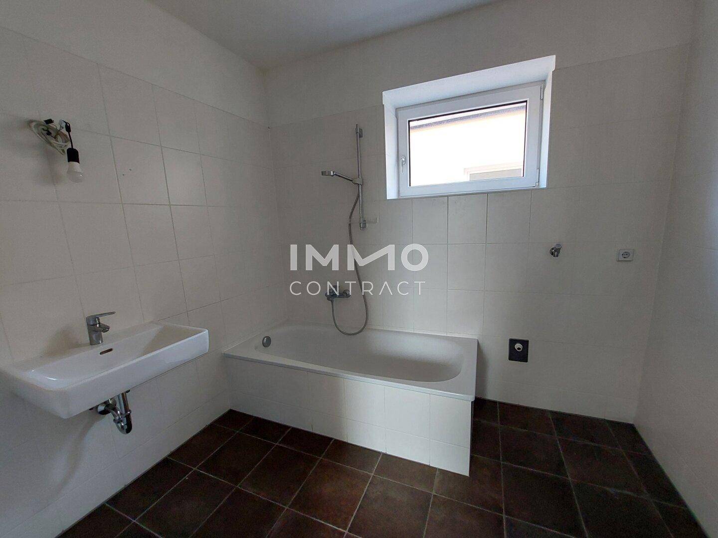 Badezimmer mit Badewanne, Waschbecken, Fliesenboden und einem Fenster für Tageslicht.
