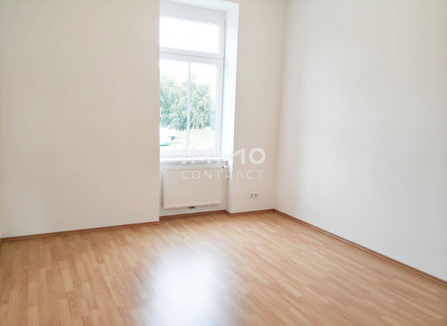 Helles Schlafzimmer mit Laminatboden und einem großen Fenster mit Blick ins Grüne.