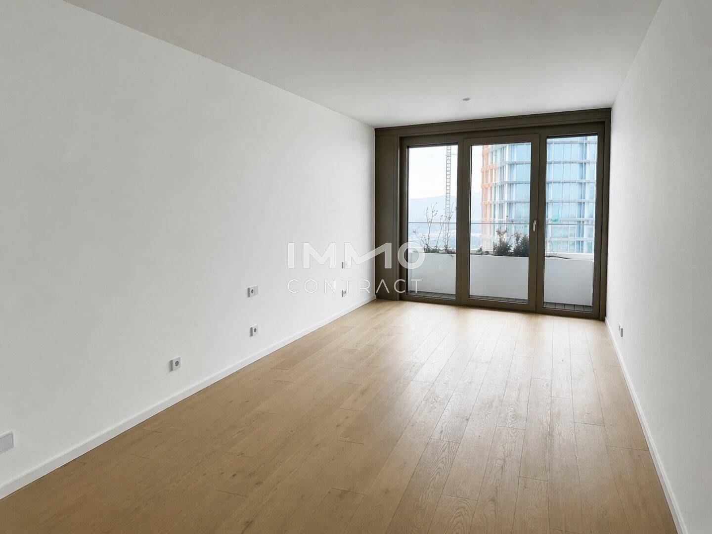 Schlafzimmer mit Holzboden und großen Fenstern, die einen Blick auf die Stadt und ein angrenzendes Gebäude ermöglichen.