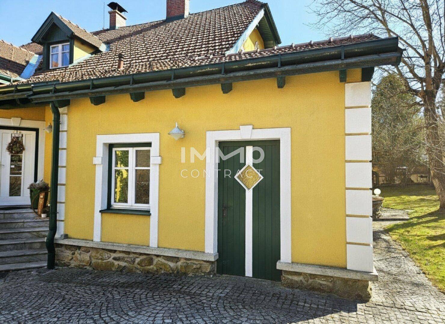 Seitenansicht des Hauses mit gelber Fassade, grünem Tor und gepflastertem Vorplatz.