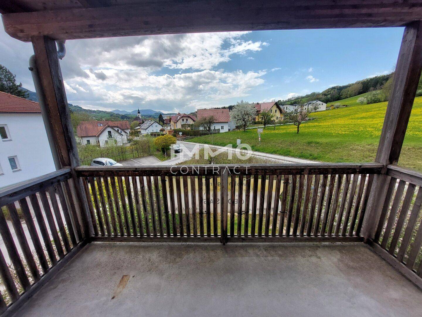 Geräumiger Holzbalkon mit weitem Blick über die grüne Landschaft und das Dorf.