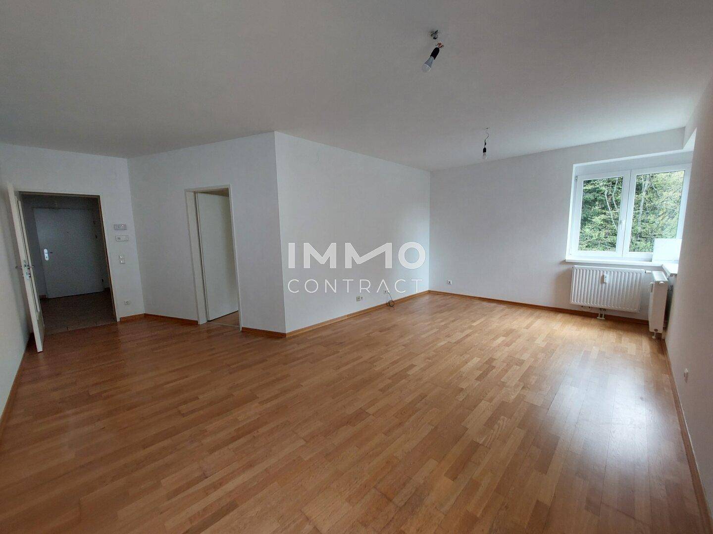Geräumiges, leeres Wohnzimmer mit hellem Parkettboden und Fenster mit Blick ins Grüne.