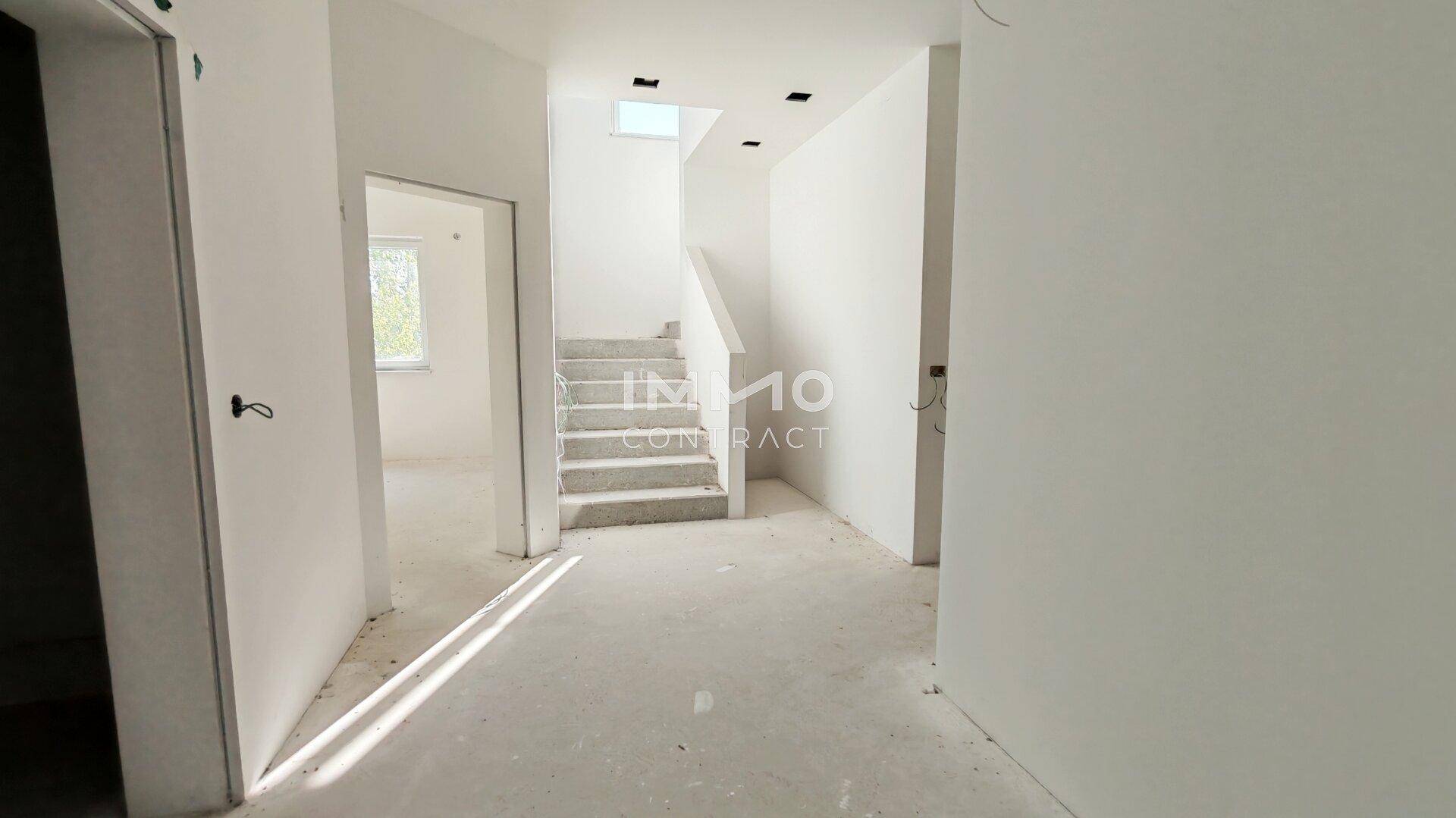 Eingangsbereich im Rohbau mit Betontreppe, weißen Wänden und natürlichem Lichteinfall.