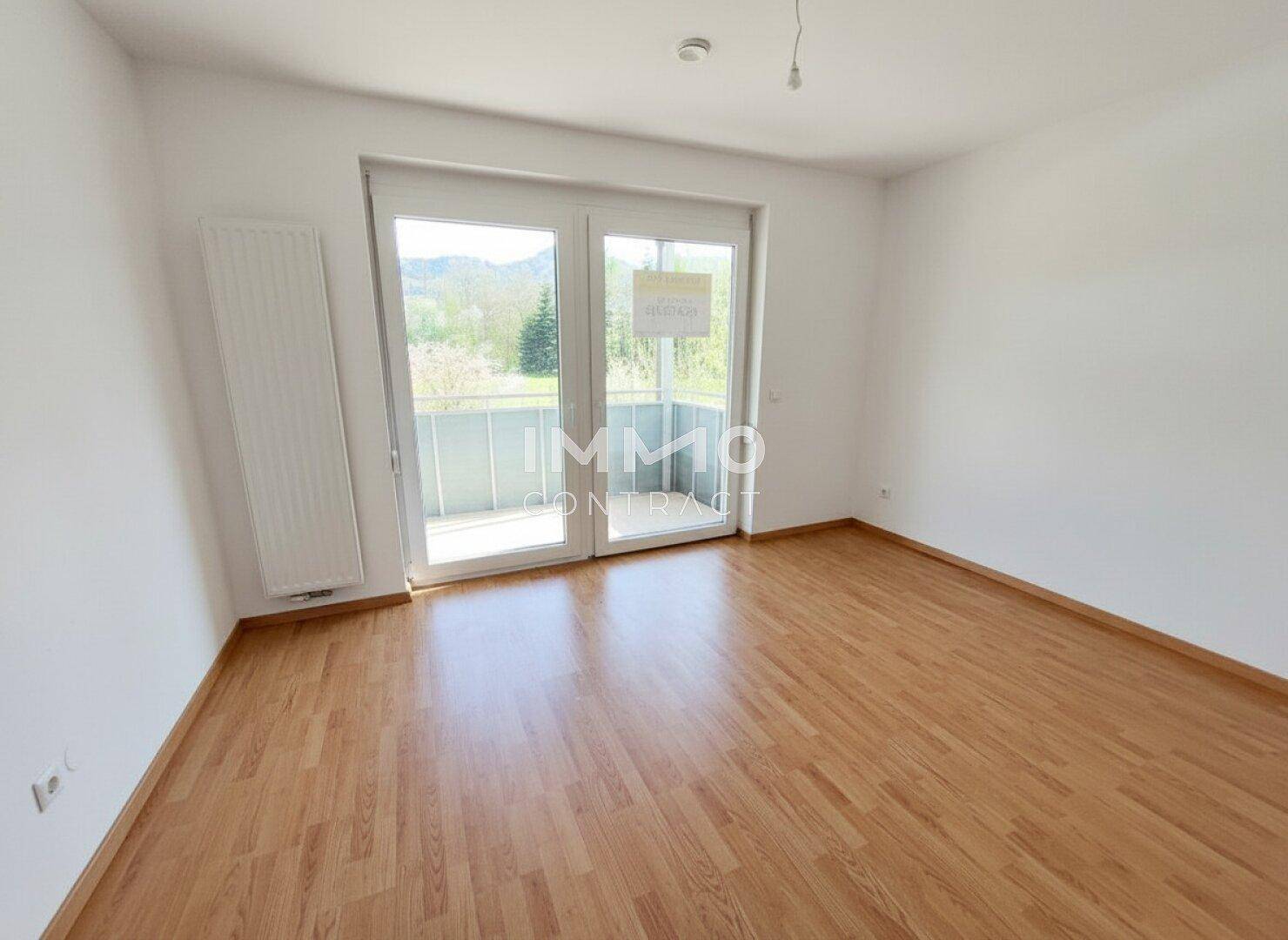 Leeres Schlafzimmer mit Laminatboden und großen Fenstern, die viel Licht hereinlassen.
