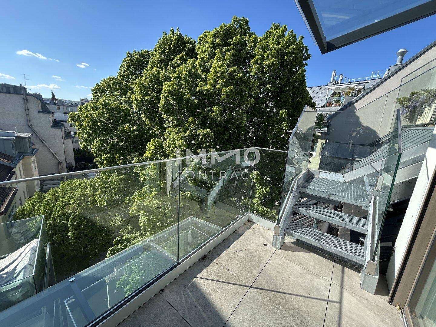 Moderne Dachterrasse mit Glasgeländer und Treppe zu einer weiteren Ebene, umgeben von Bäumen.