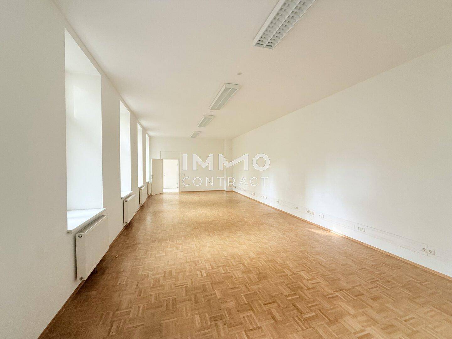 Langer, heller Raum mit Parkettboden und einer Reihe von Fenstern, ideal für ein Großraumbüro oder mehrere Arbeitsbereiche.