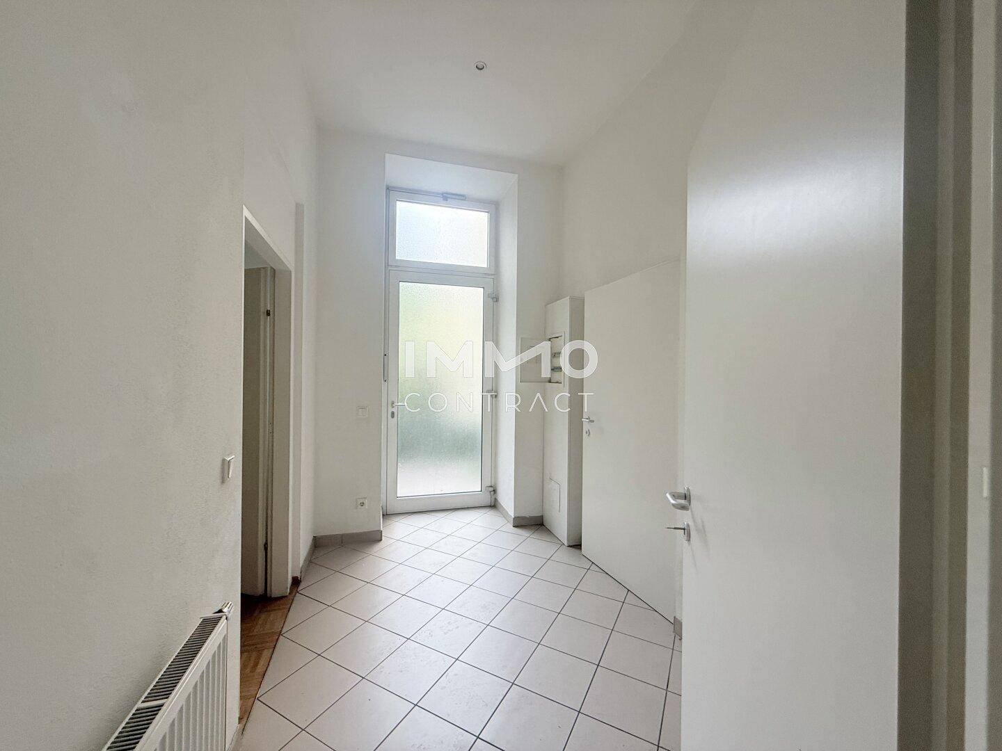 Heller Flur mit Fliesenboden und einer Tür, die ins Freie führt, sowie einem Heizkörper an der Wand.