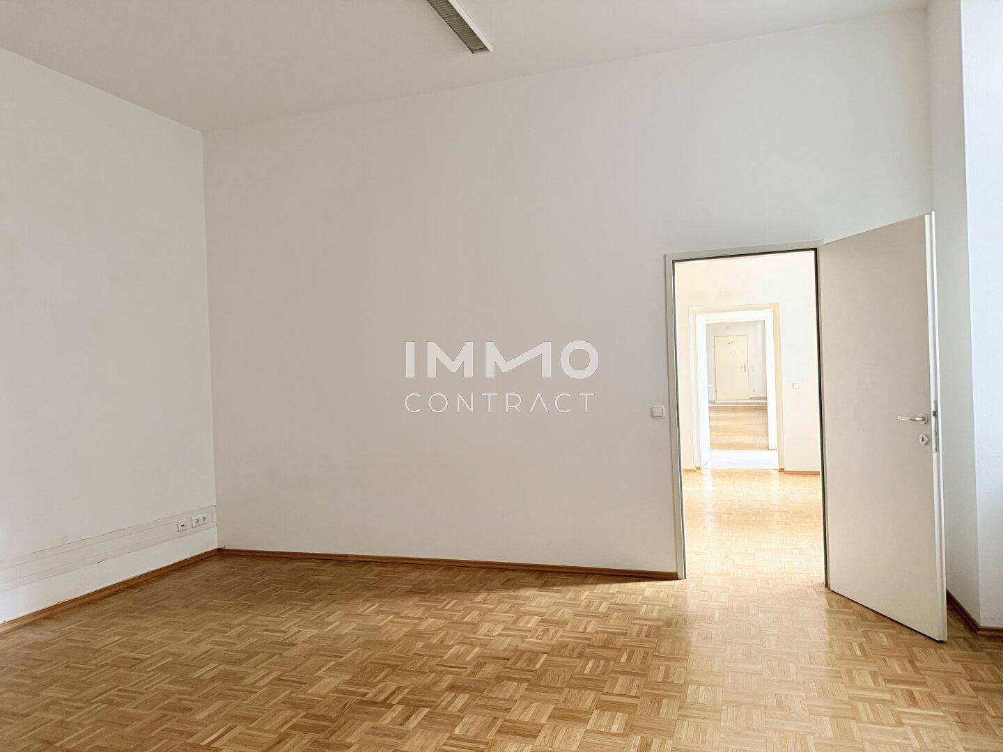 Geräumiger Büroraum mit Parkettboden und offener Tür, die den Blick in weitere helle Räume freigibt.