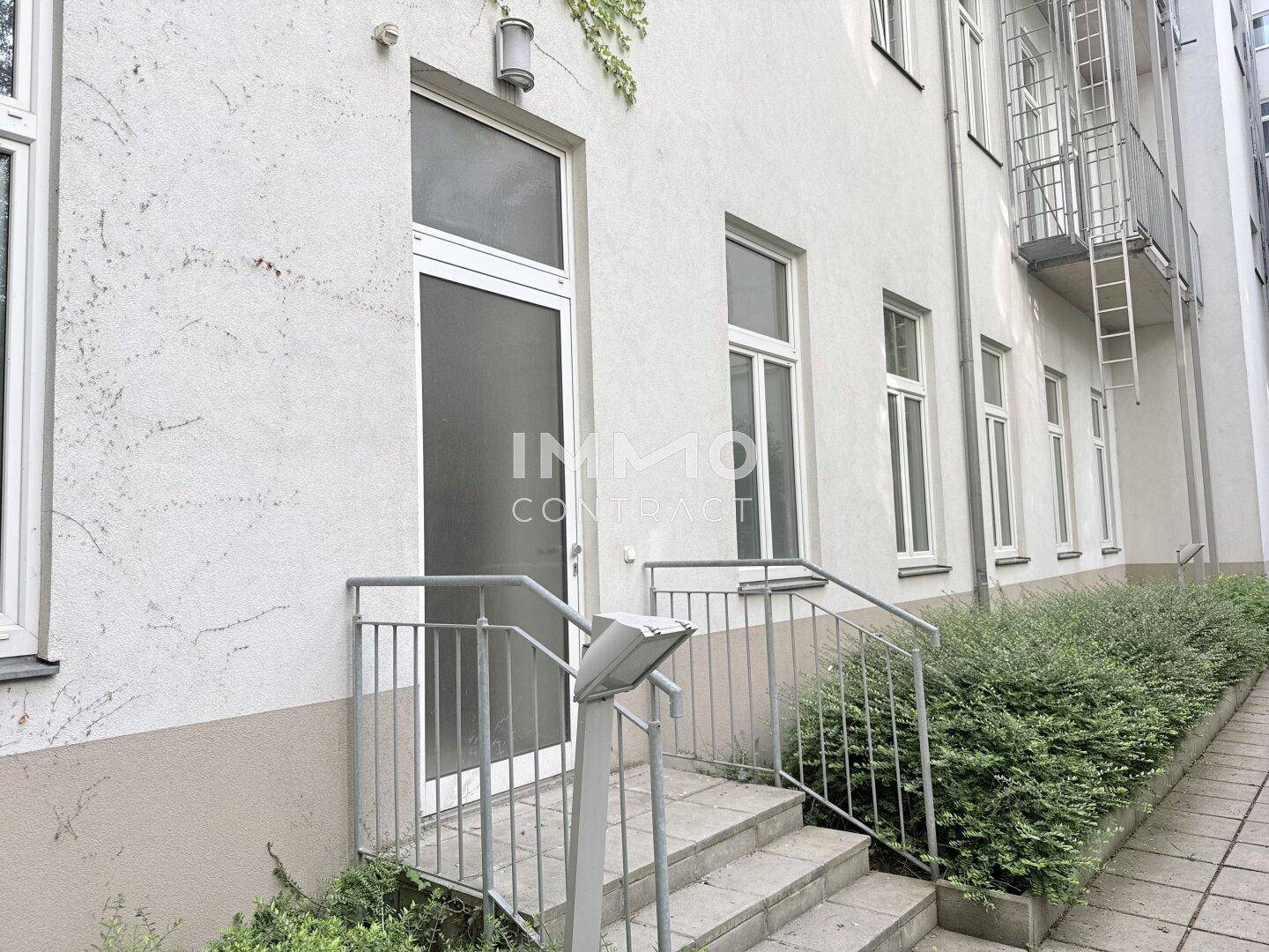 Eingangsbereich des Gebäudes mit Treppe, Geländer und gepflegten Büschen entlang der Fassade, die zum Eingang führen.
