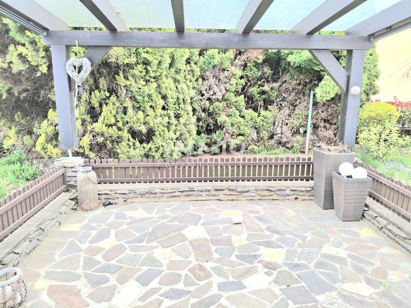 Geräumige gepflasterte Terrasse unter einer Pergola, umgeben von hohen Hecken.