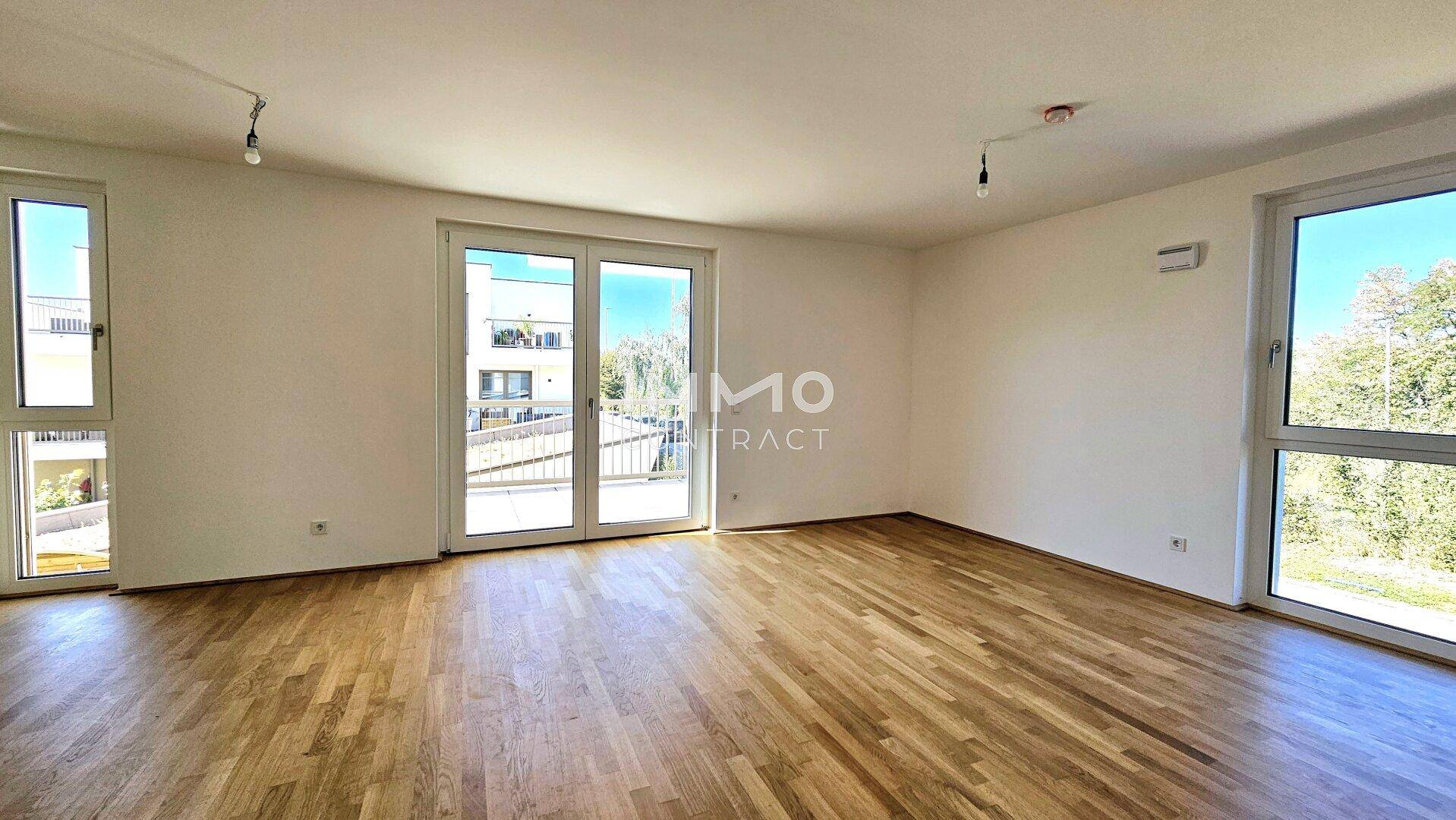 Geräumiges Wohnzimmer mit hellem Parkettboden und großen Fenstern, die viel Tageslicht hereinlassen.