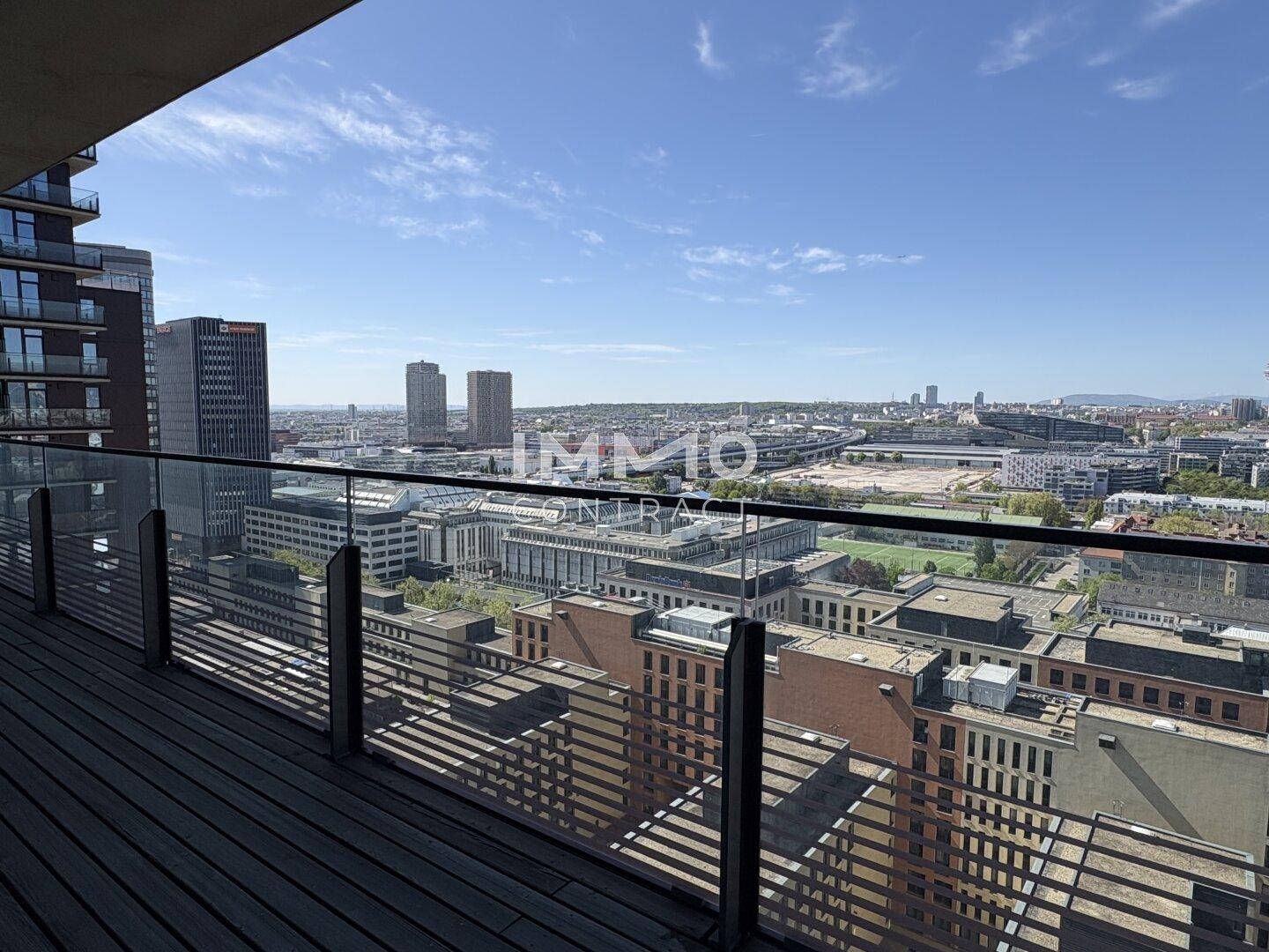 Geräumiger Balkon mit Holzboden, Glasgeländer und weitem Blick über die Stadt.