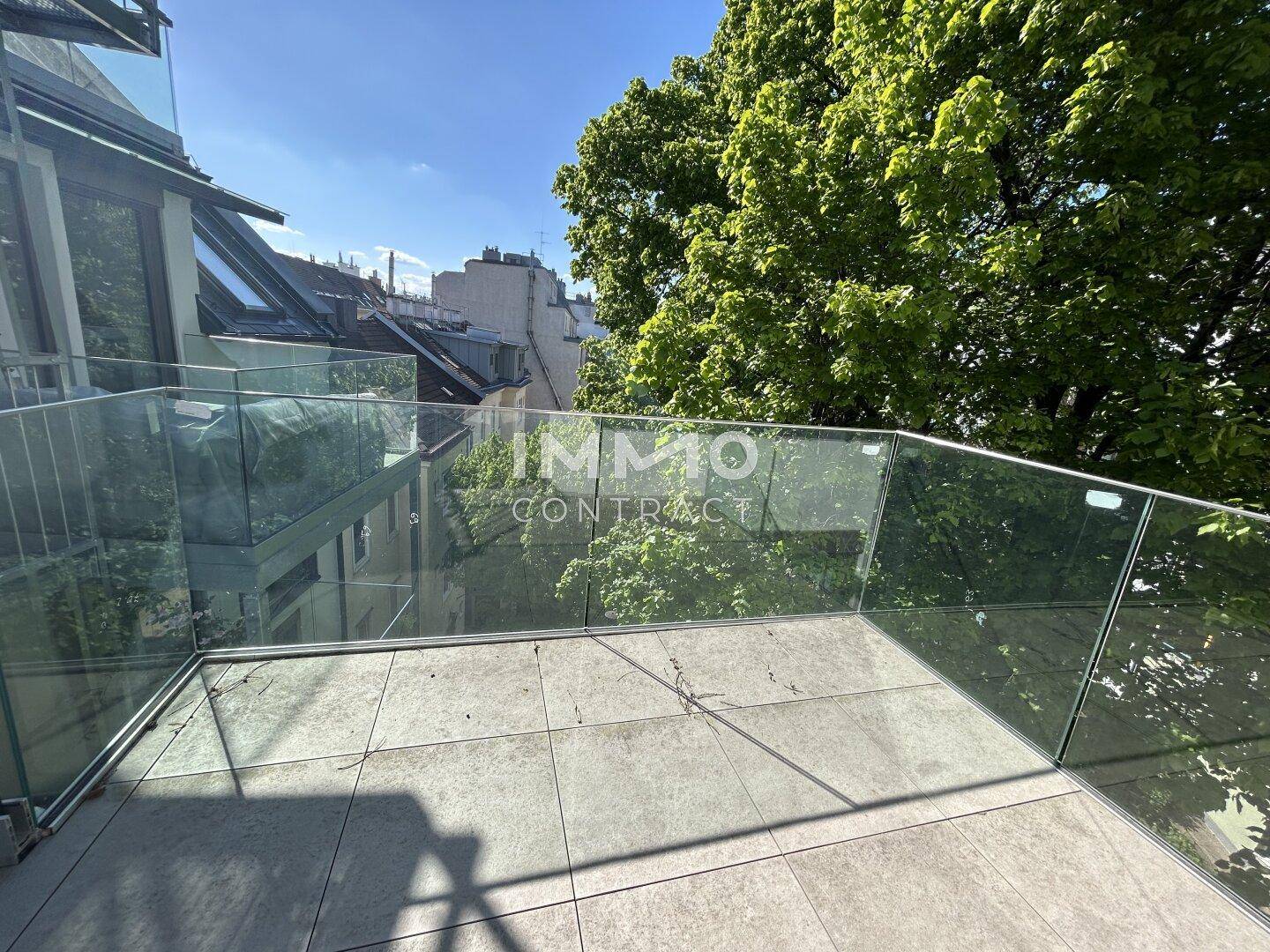 Kleine Terrasse mit modernem Glasgeländer und Blick auf grüne Bäume.