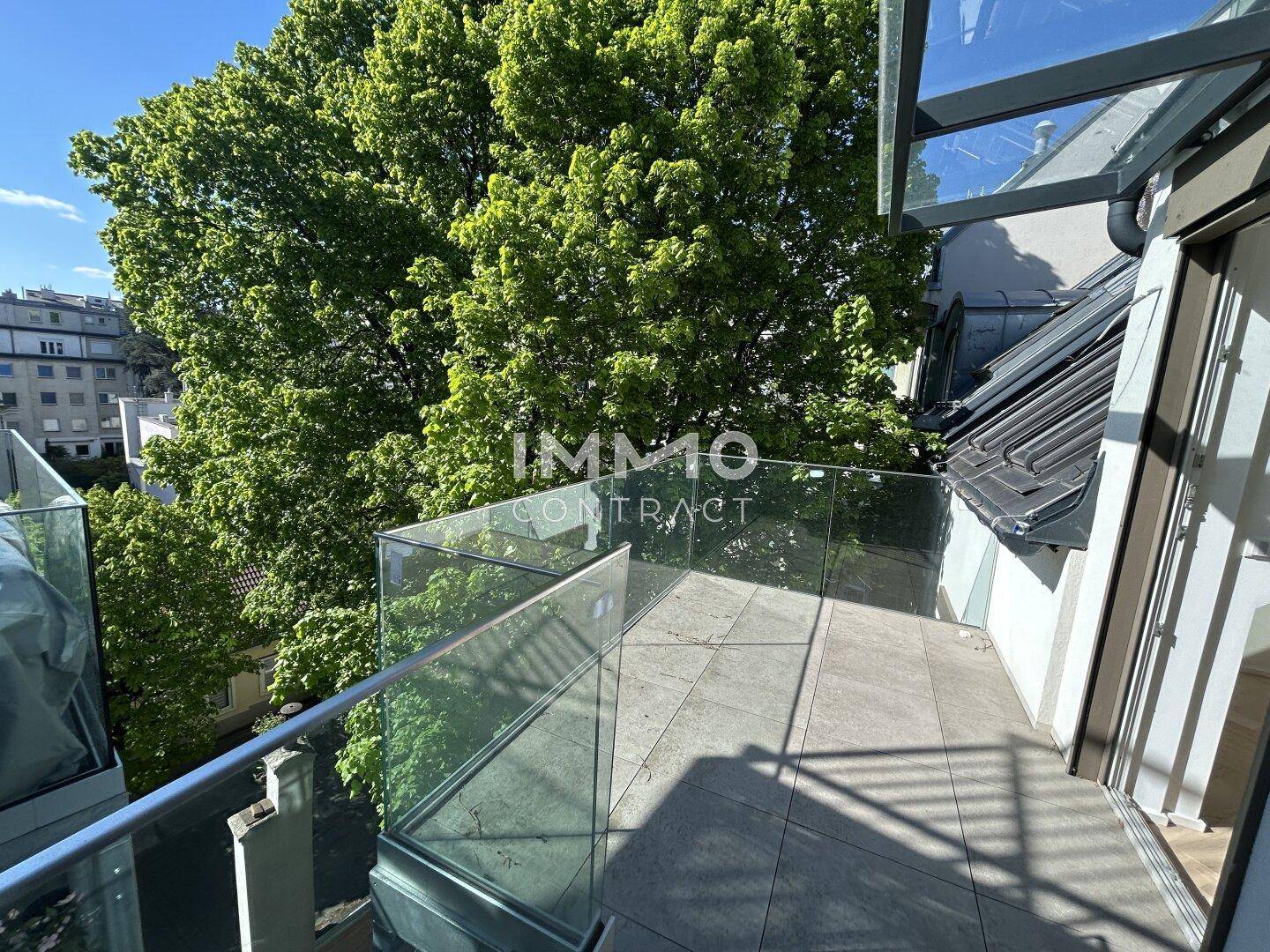 Sonniger Terrassenbereich mit Glasgeländer und üppigem Baumbestand in der Umgebung.