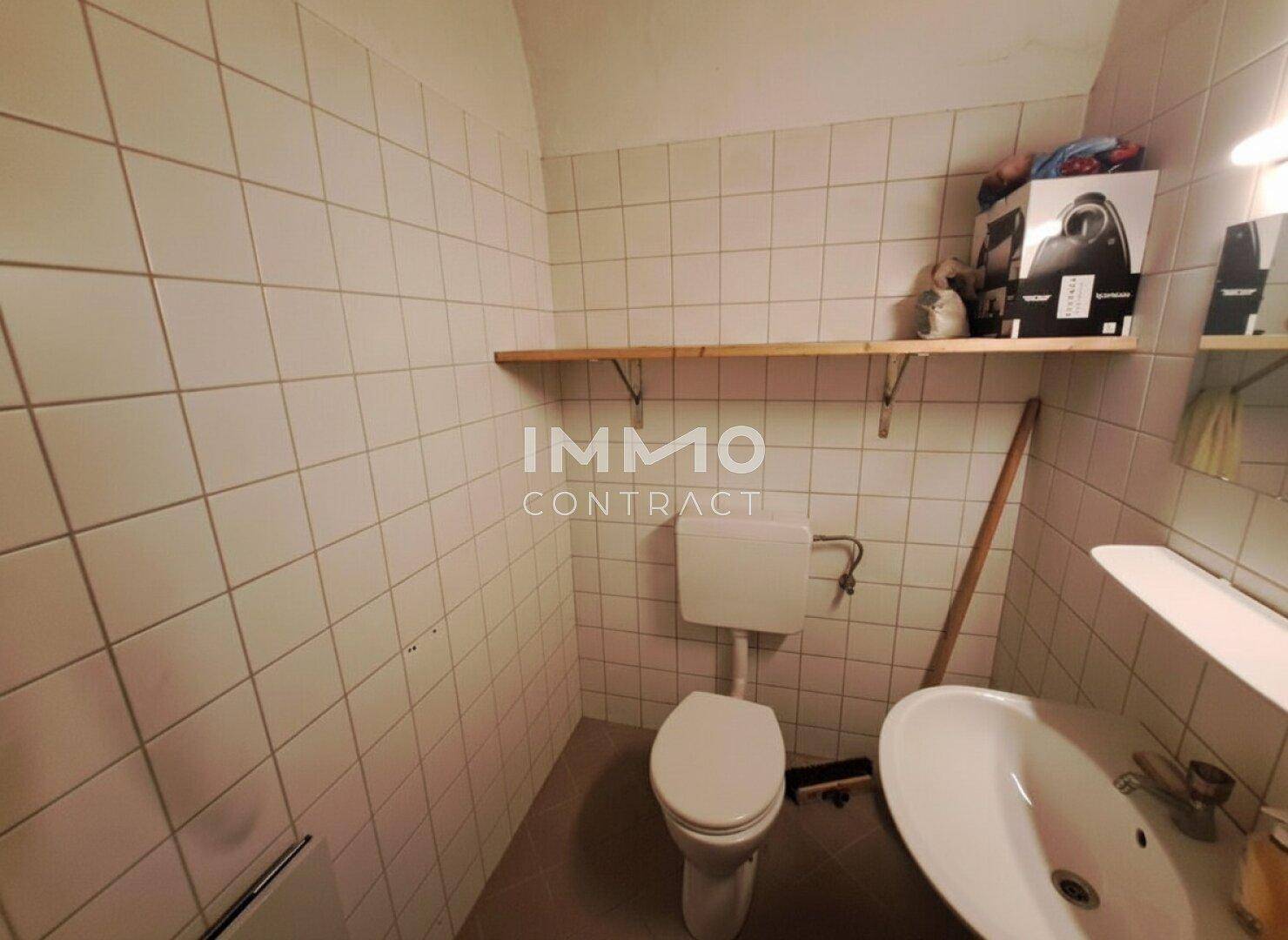 Einfache Toilette mit weißen Fliesen an den Wänden, einem WC und einem Waschbecken.