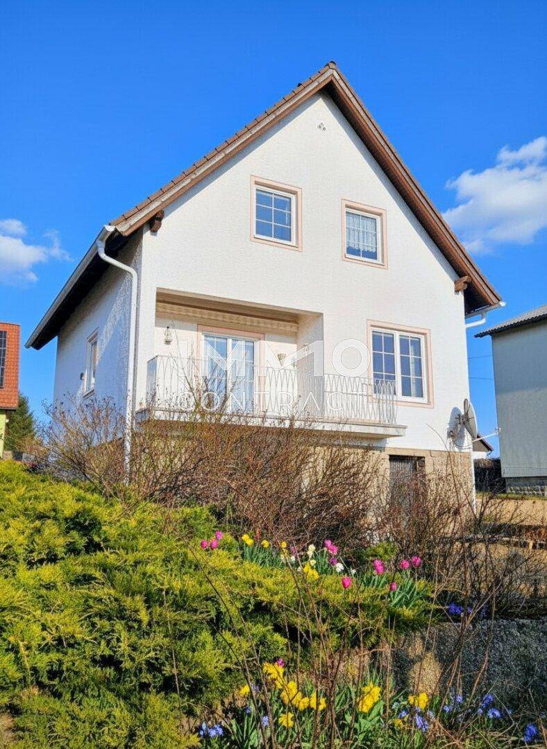 Vorderansicht des weißen Hauses mit blühenden Blumen und einem Balkon unter blauem Himmel.