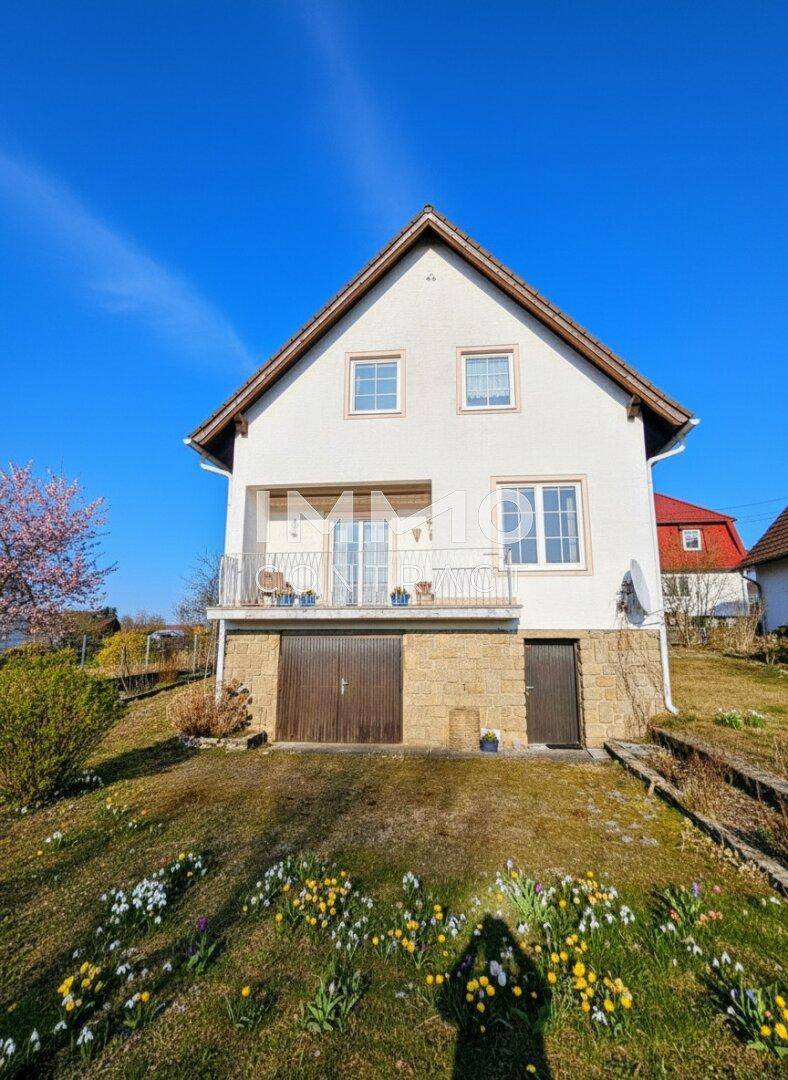 Vorderansicht des Hauses mit Garage, Balkon und blühender Wiese im Frühling.