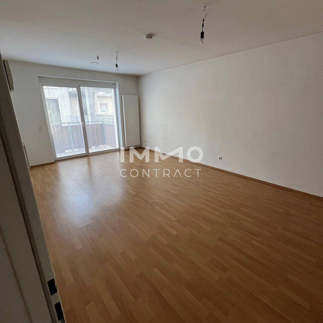 Leeres Wohnzimmer mit hellem Laminatboden, weißen Wänden und großem Fenster mit Balkonzugang.