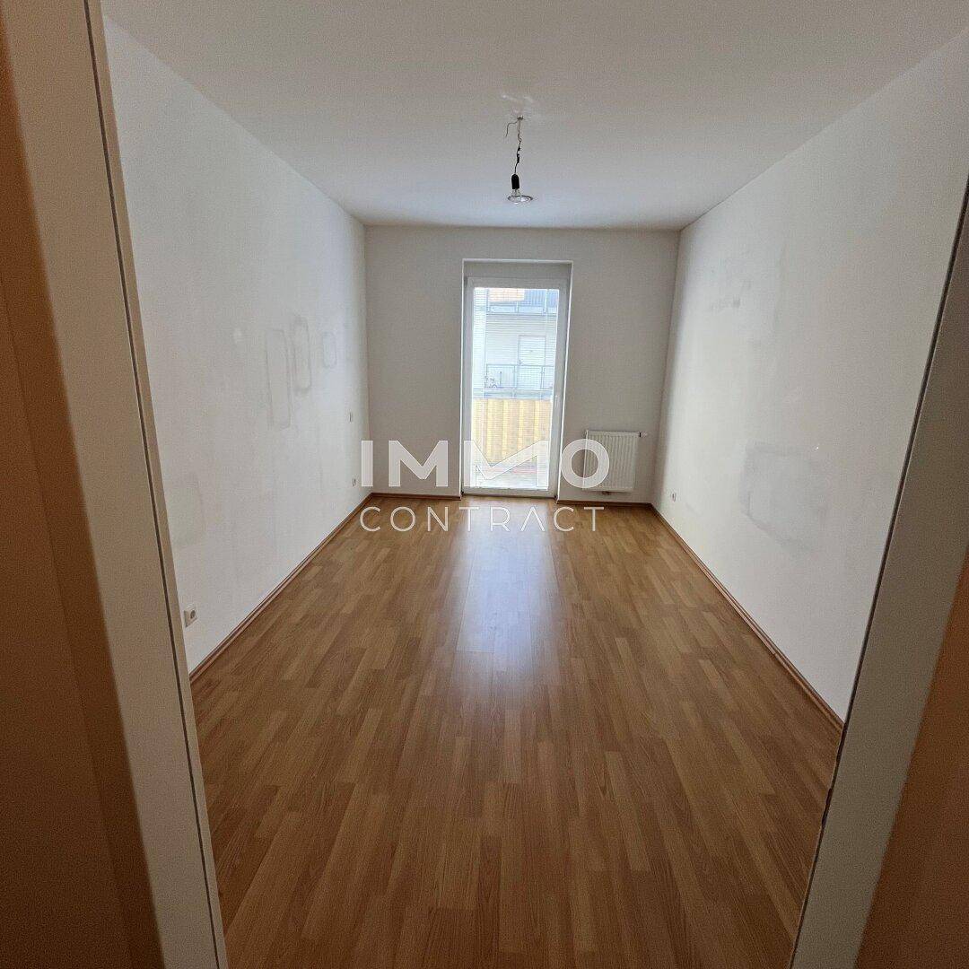 Helles, leeres Schlafzimmer mit Laminatboden und Blick nach draußen durch das Fenster.