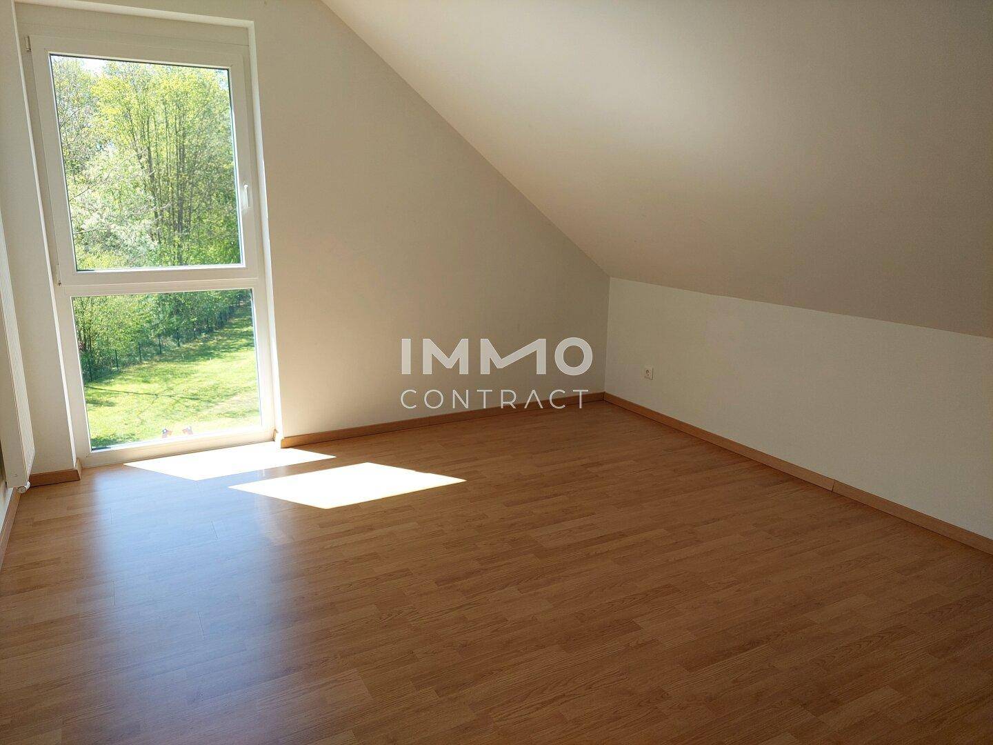 Heller, leerer Raum mit Holzboden, großem Fenster und Blick ins Grüne, ideal als Schlafzimmer.