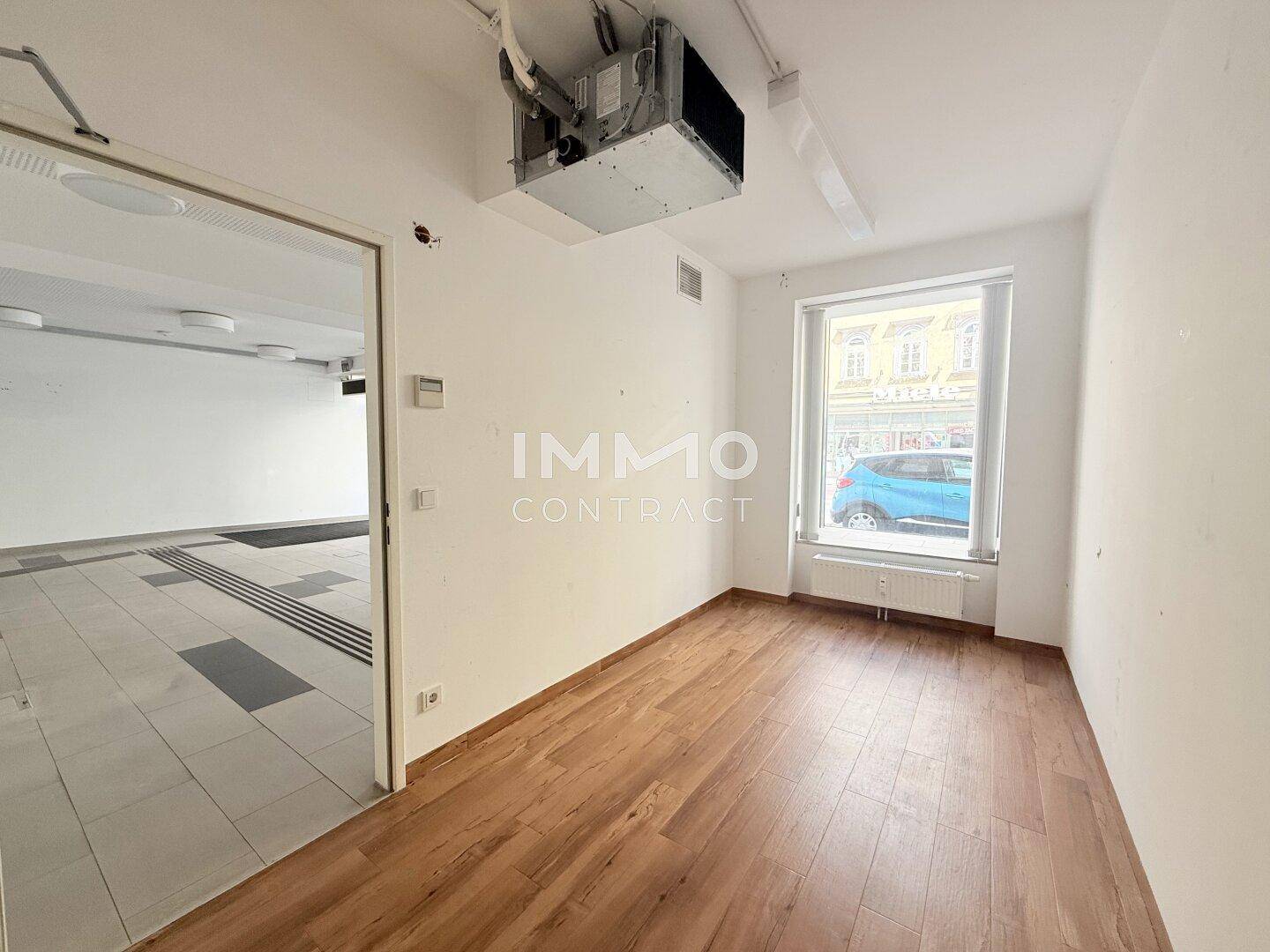 Kleineres Büro mit Holzboden und Fenster zur Straße, verbunden mit dem Hauptraum, hell und funktional.