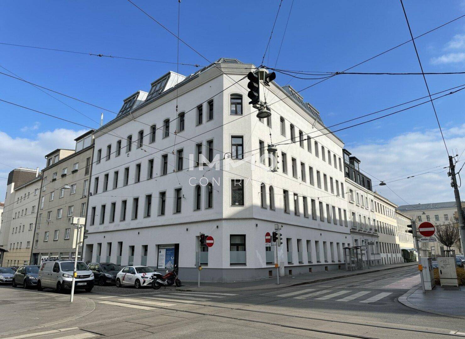 Modernes Eckgebäude mit weißer Fassade und vielen Fenstern in urbaner Umgebung.