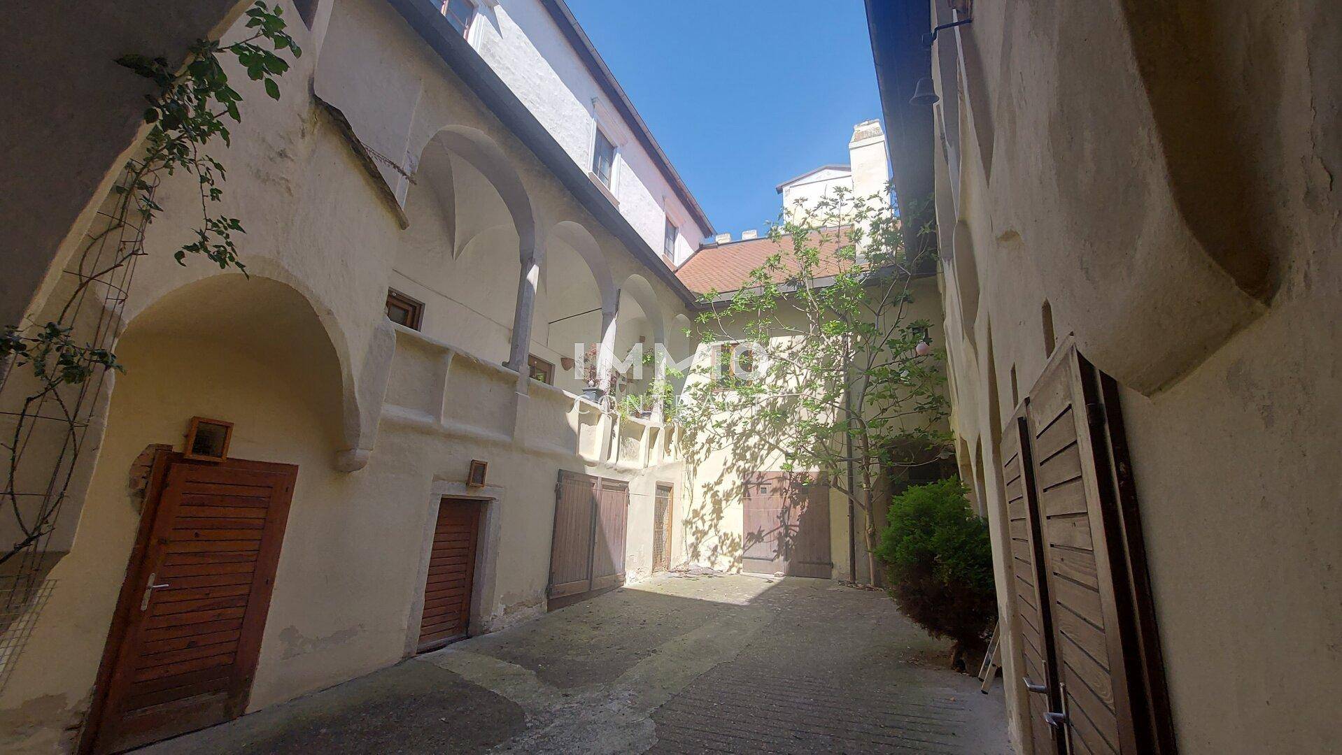 Sonniger Innenhof mit Arkadengängen, alten Holztüren und einem Baum.
