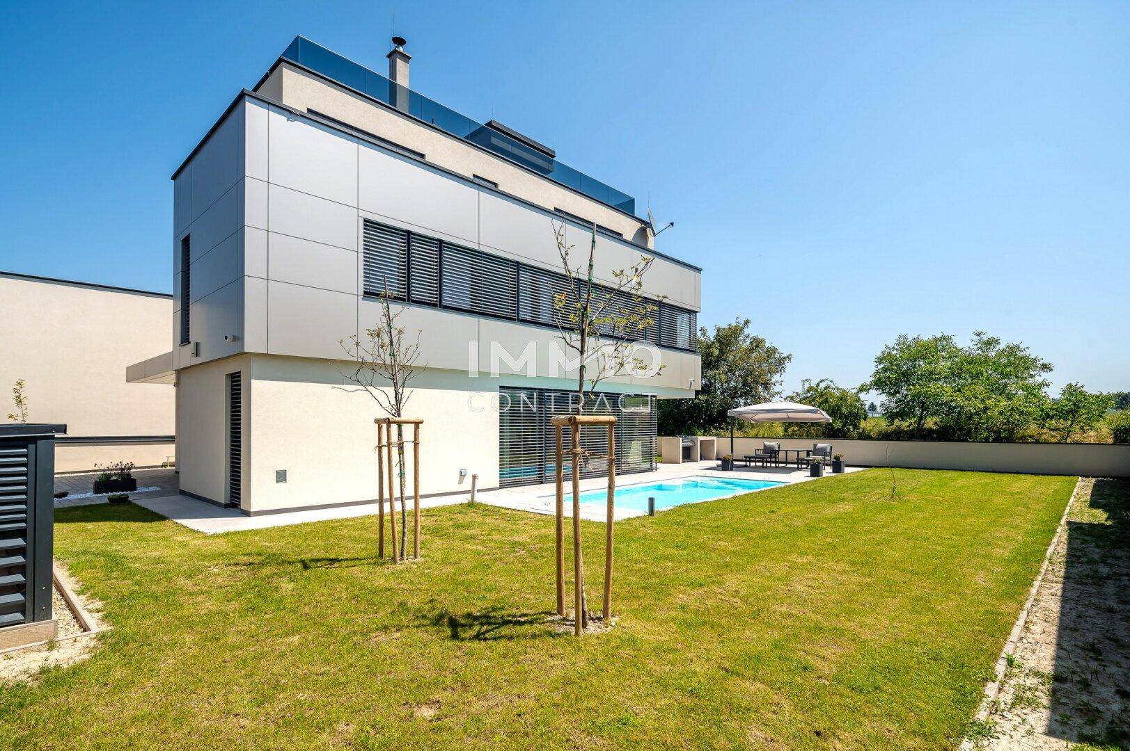 Seitenansicht der modernen Villa mit Poolbereich und weitläufigem Rasen, umgeben von jungen Bäumen.