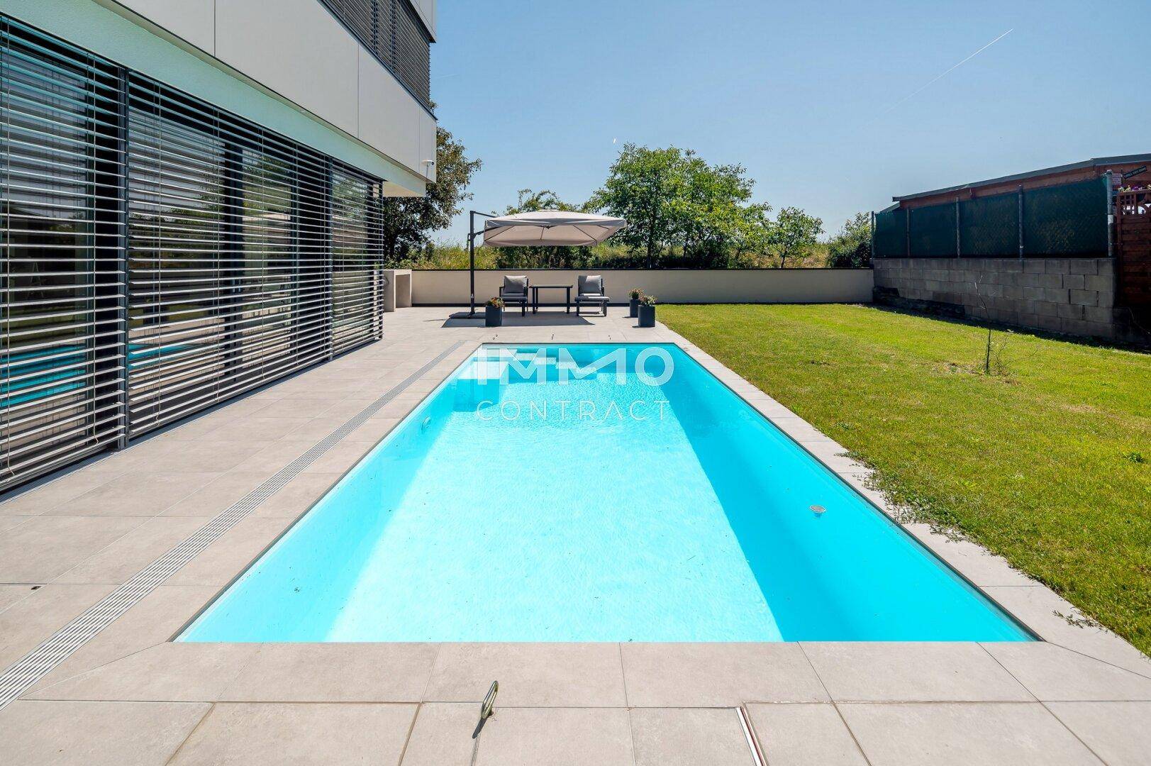 Langer Swimmingpool neben der Hausfassade, umgeben von einer hellen Terrasse und Rasenfläche.