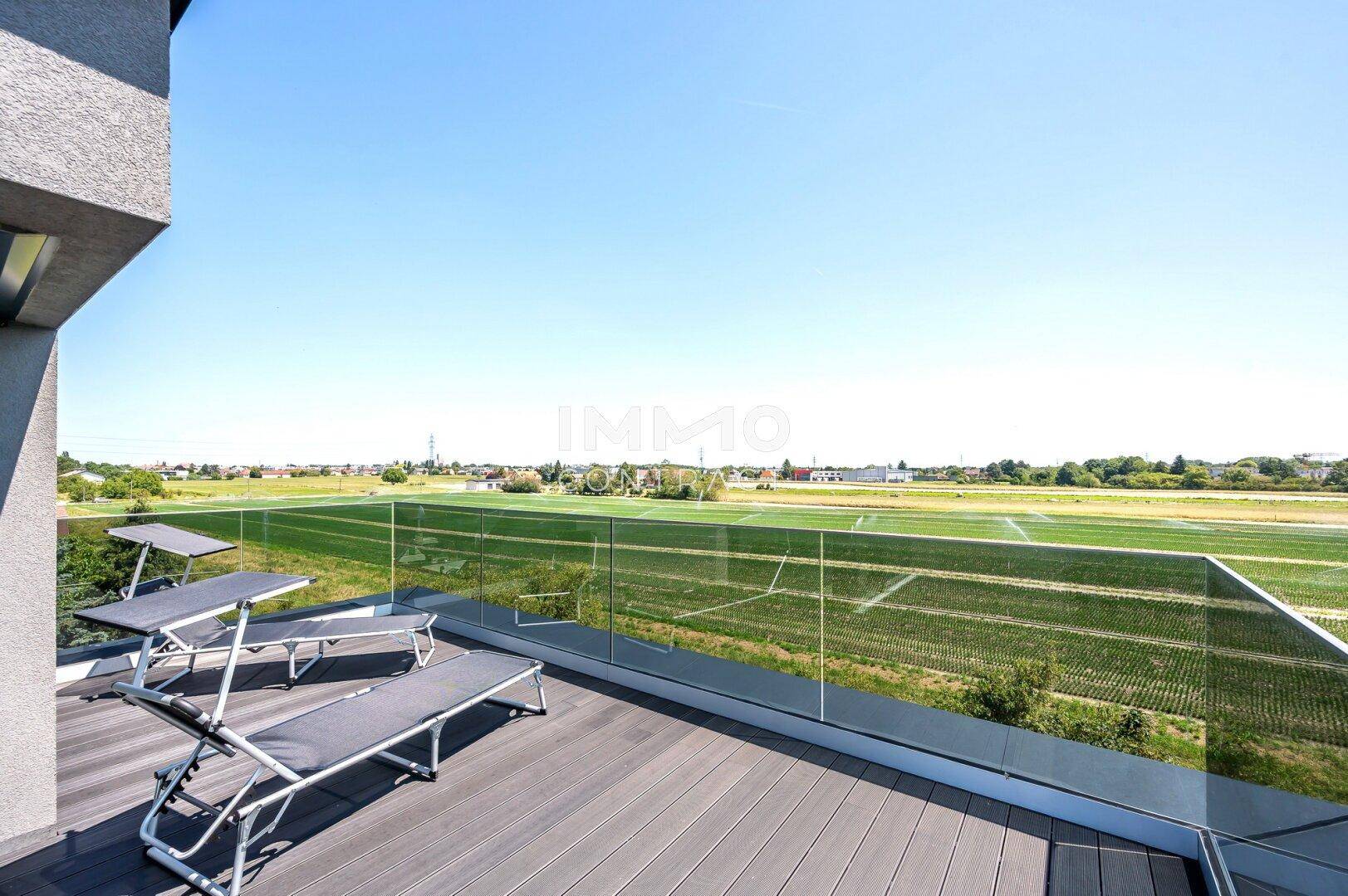 Sonniger Dachterrasse mit Liegestühlen und Glasgeländer, bietet einen weiten Blick über Felder.