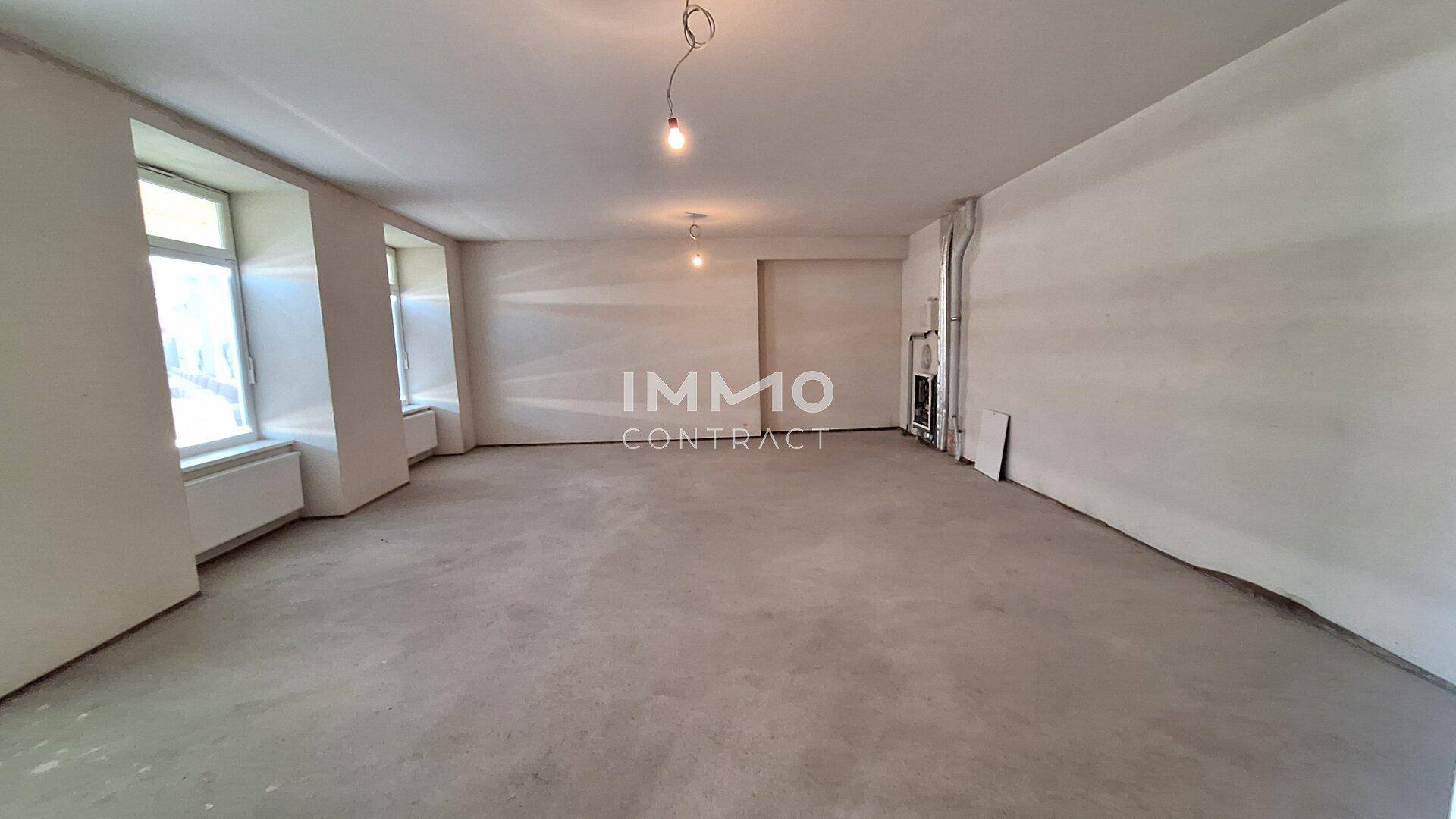 Weiterer Blick in den großen, leeren Raum mit mehreren Fenstern, Betonboden und viel Tageslicht.