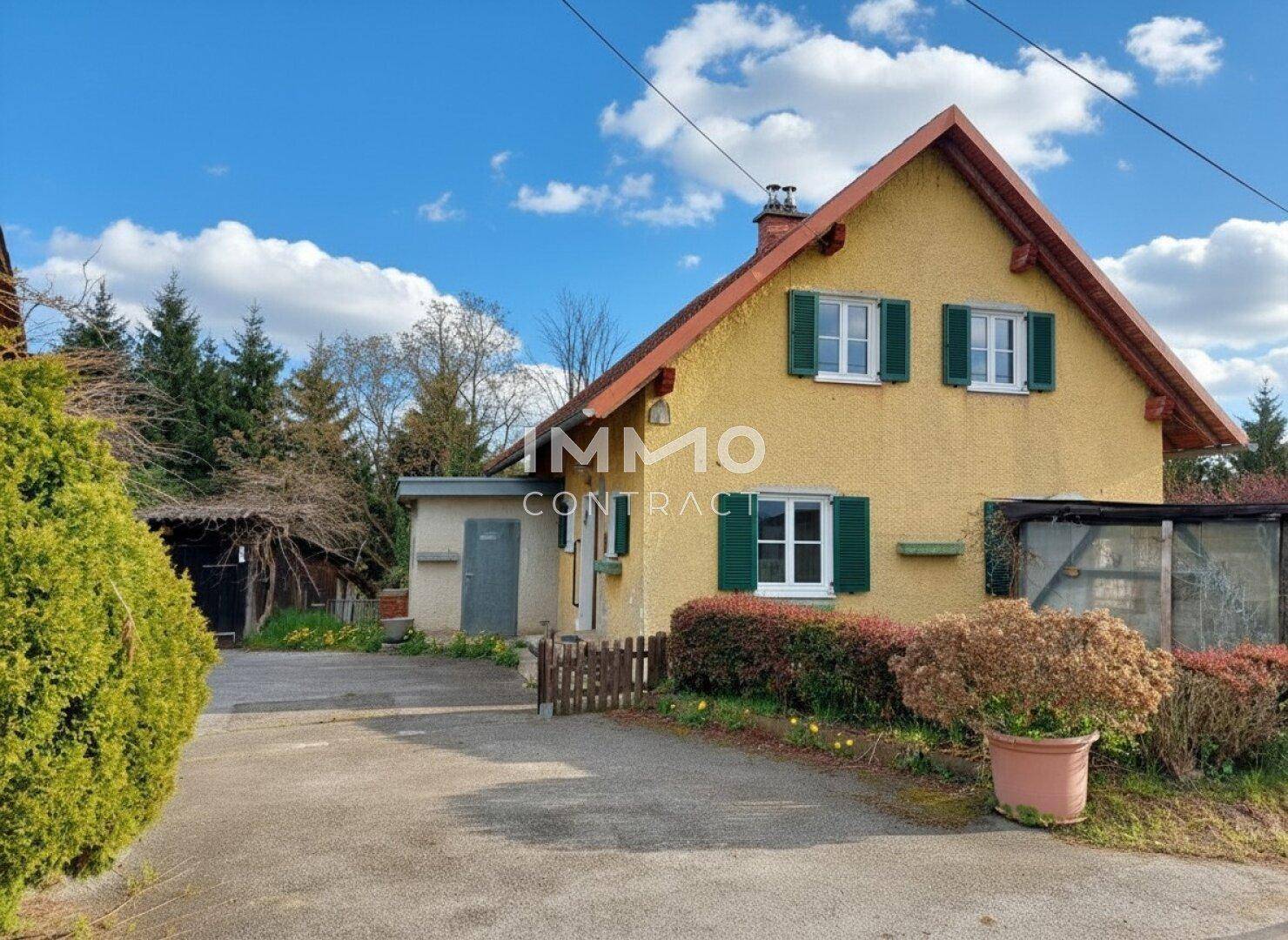 Gelbes Landhaus mit grünen Fensterläden und rotem Dach in ländlicher Umgebung.