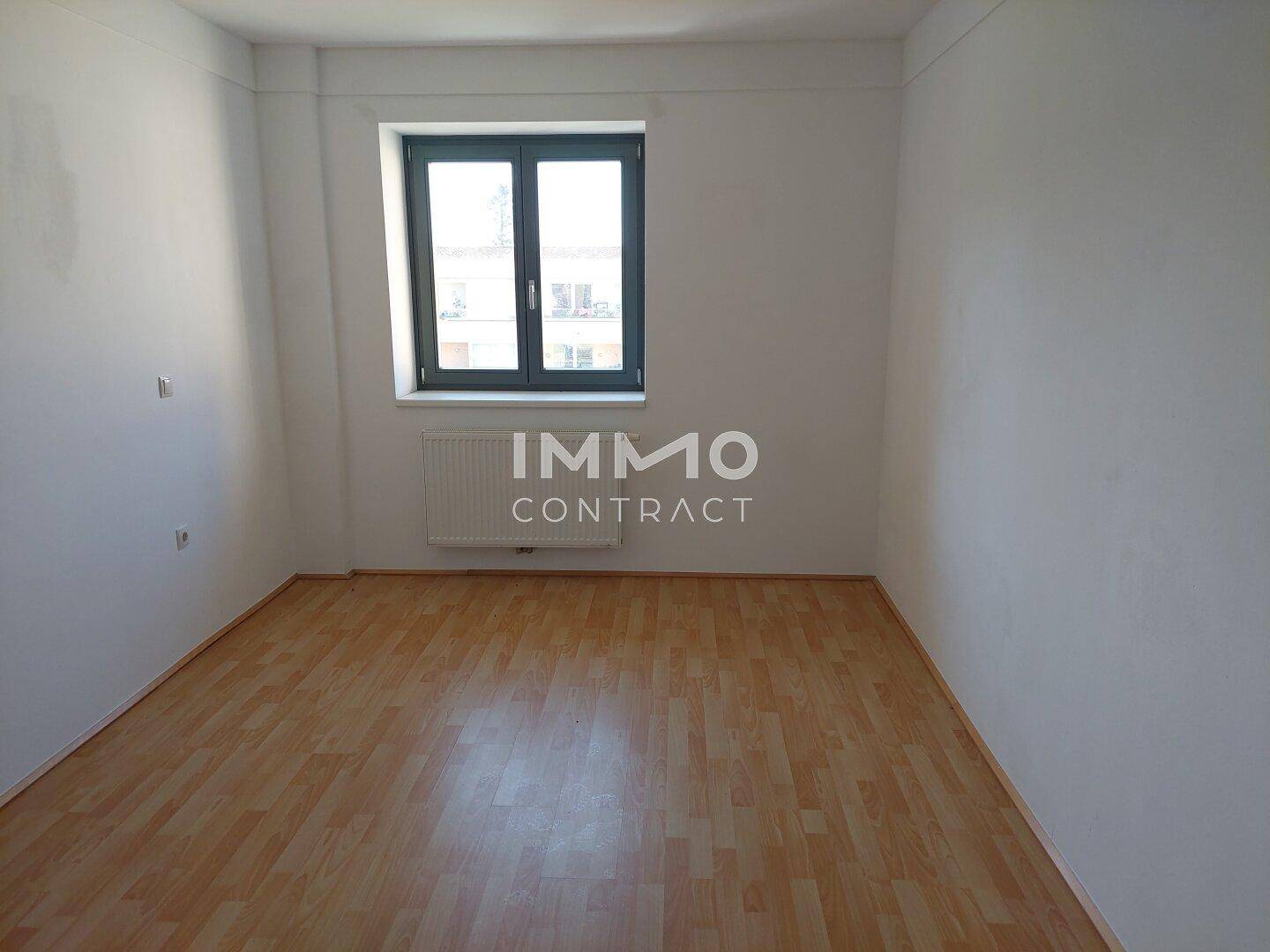 Leerer Raum mit hellem Laminatboden, Fenster und Heizkörper, vielseitig nutzbar.