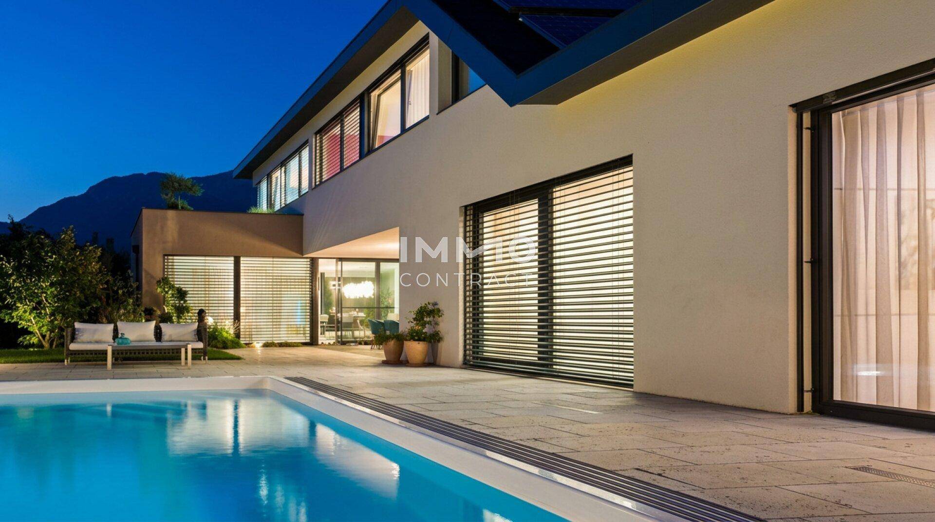Modernes Haus bei Abenddämmerung mit beleuchtetem Pool und großen Fenstern mit Jalousien.