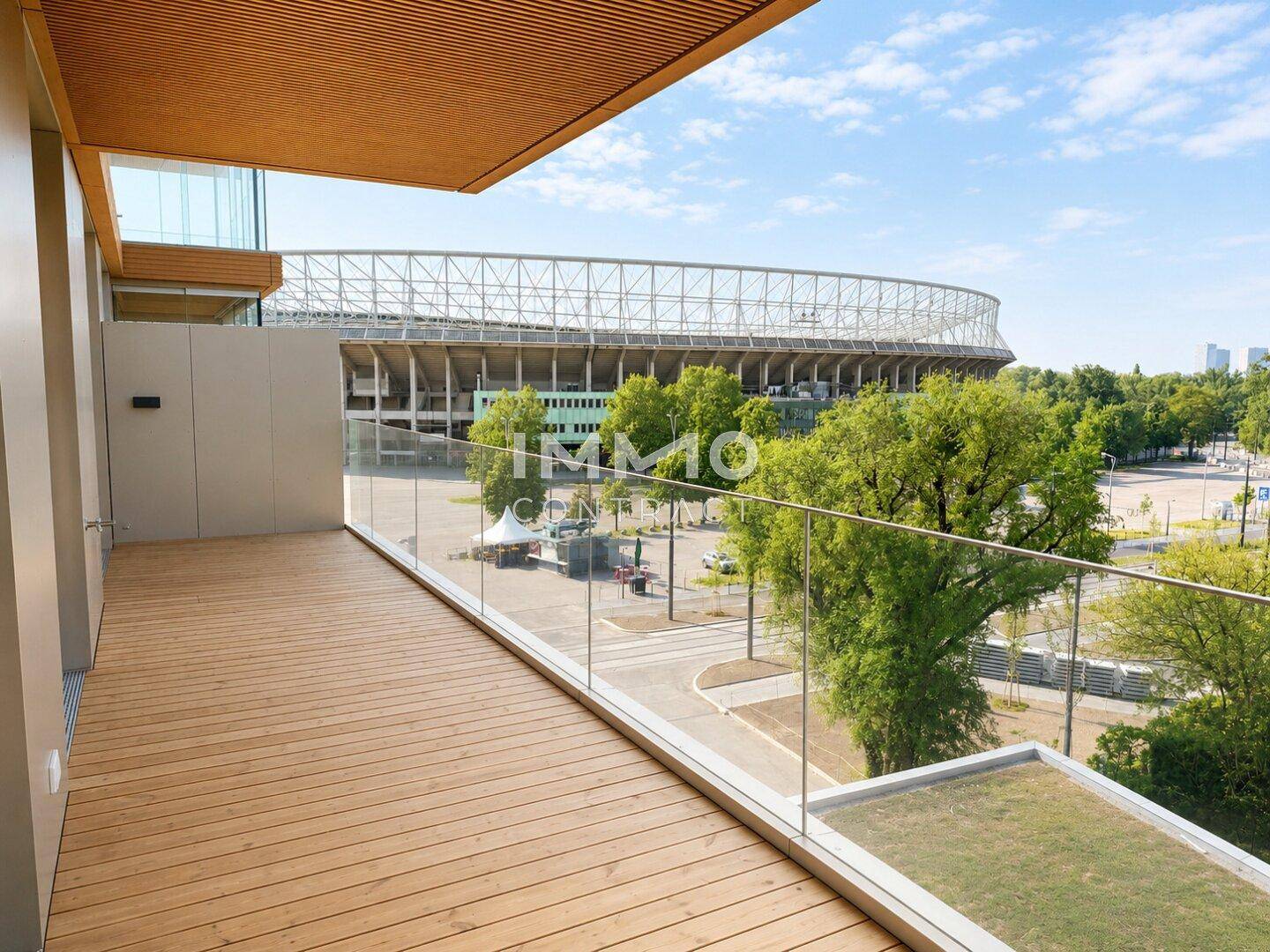 Balkon mit Holzboden, Glasgeländer und Blick auf ein nahegelegenes Sportstadion.