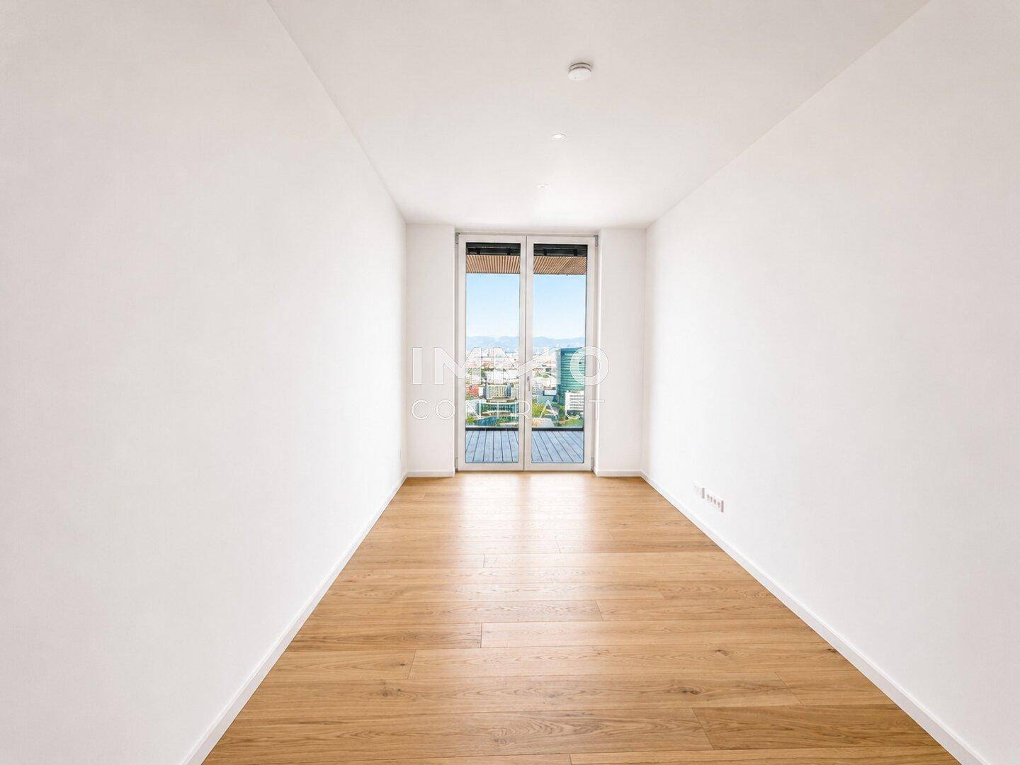 Helles, leeres Zimmer mit Eichenparkett und Zugang zu einem Balkon mit Stadtblick.