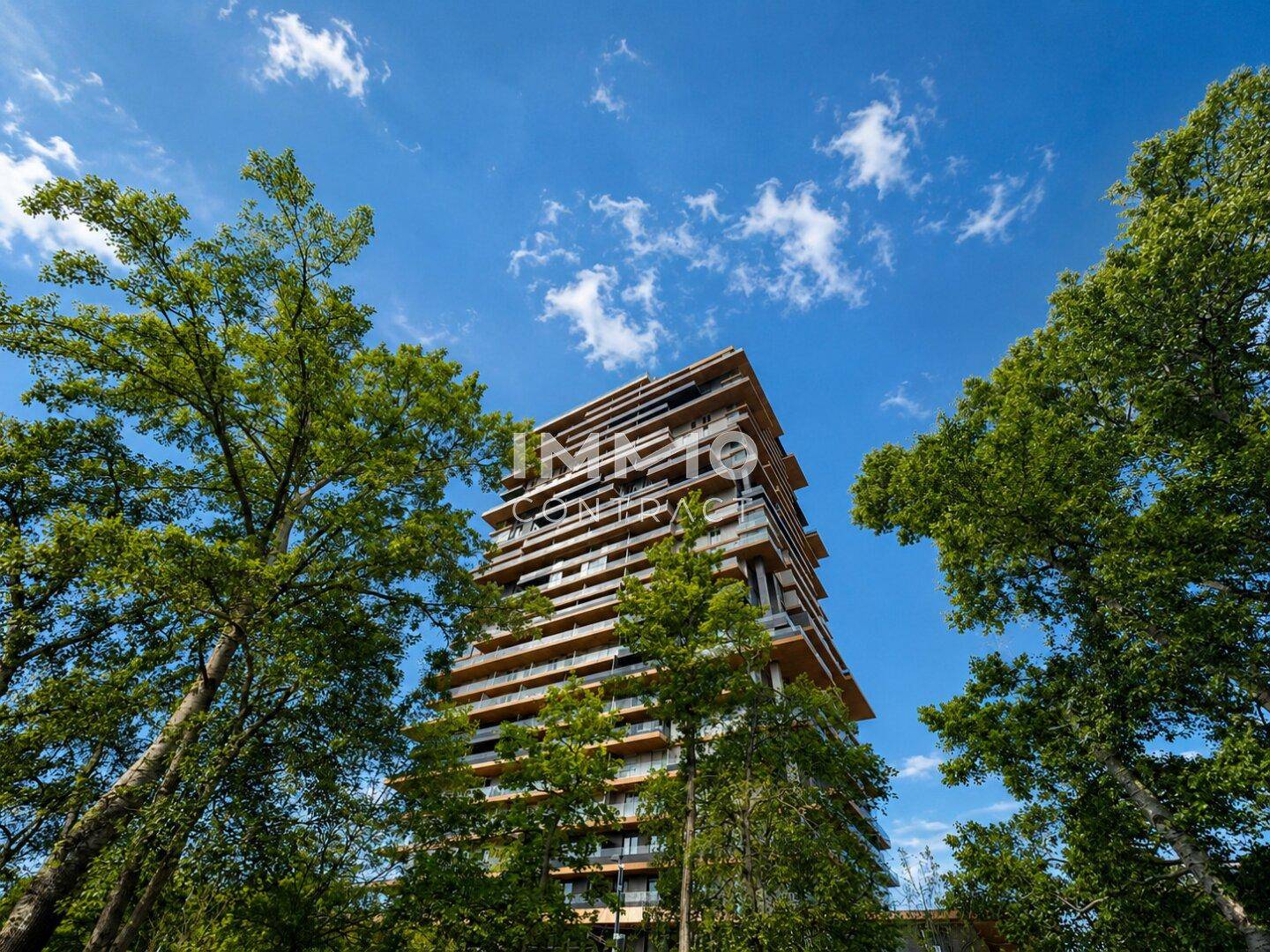 Beeindruckendes modernes Hochhaus mit vielen Balkonen, umgeben von hohen Bäumen unter blauem Himmel.