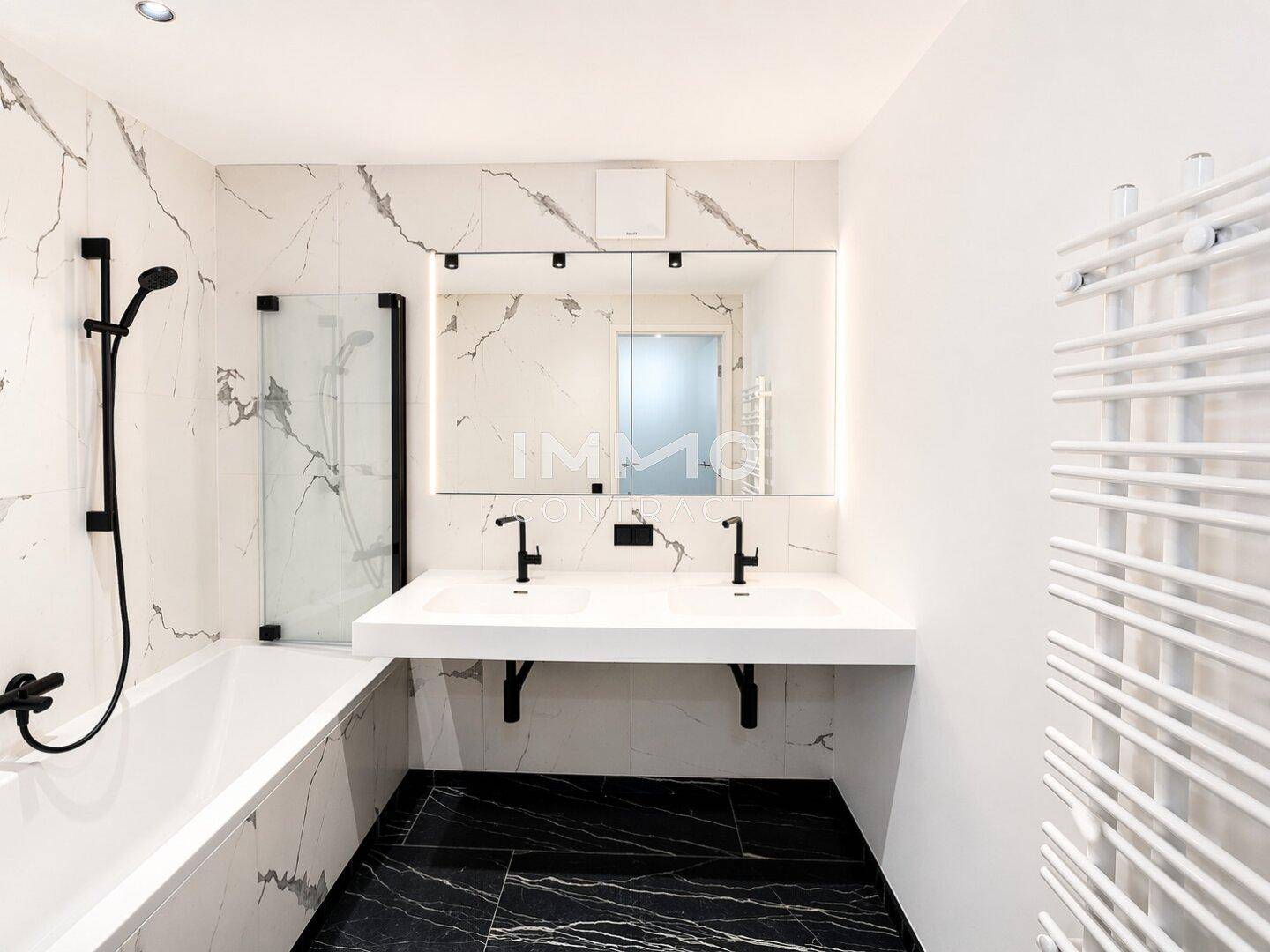 Modernes Badezimmer mit Marmoroptik-Fliesen, Doppelwaschbecken und Badewanne mit Duschfunktion.